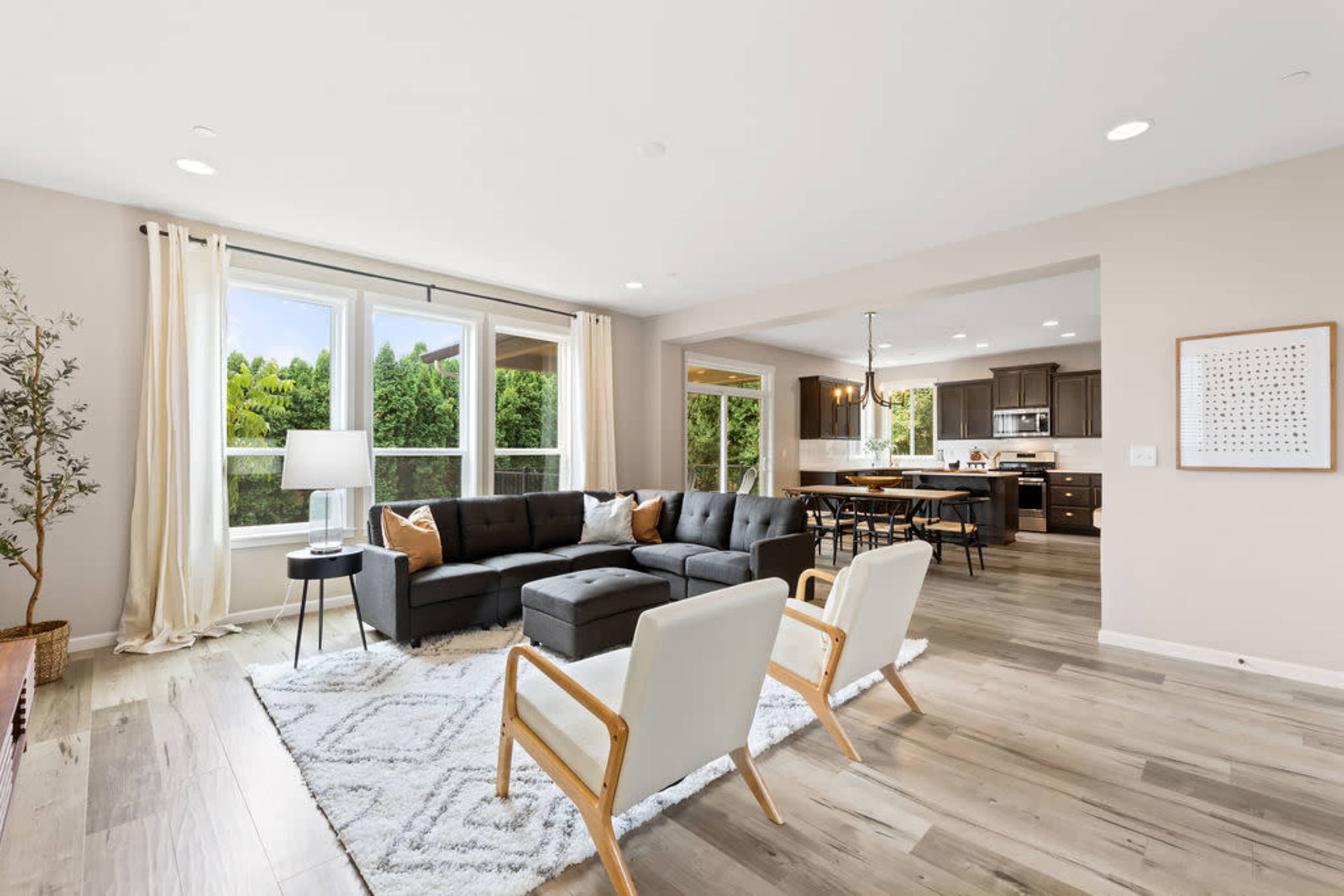 A modern living room with a gray sectional sofa, two light-colored chairs, a table lamp, and large windows overlooking greenery.