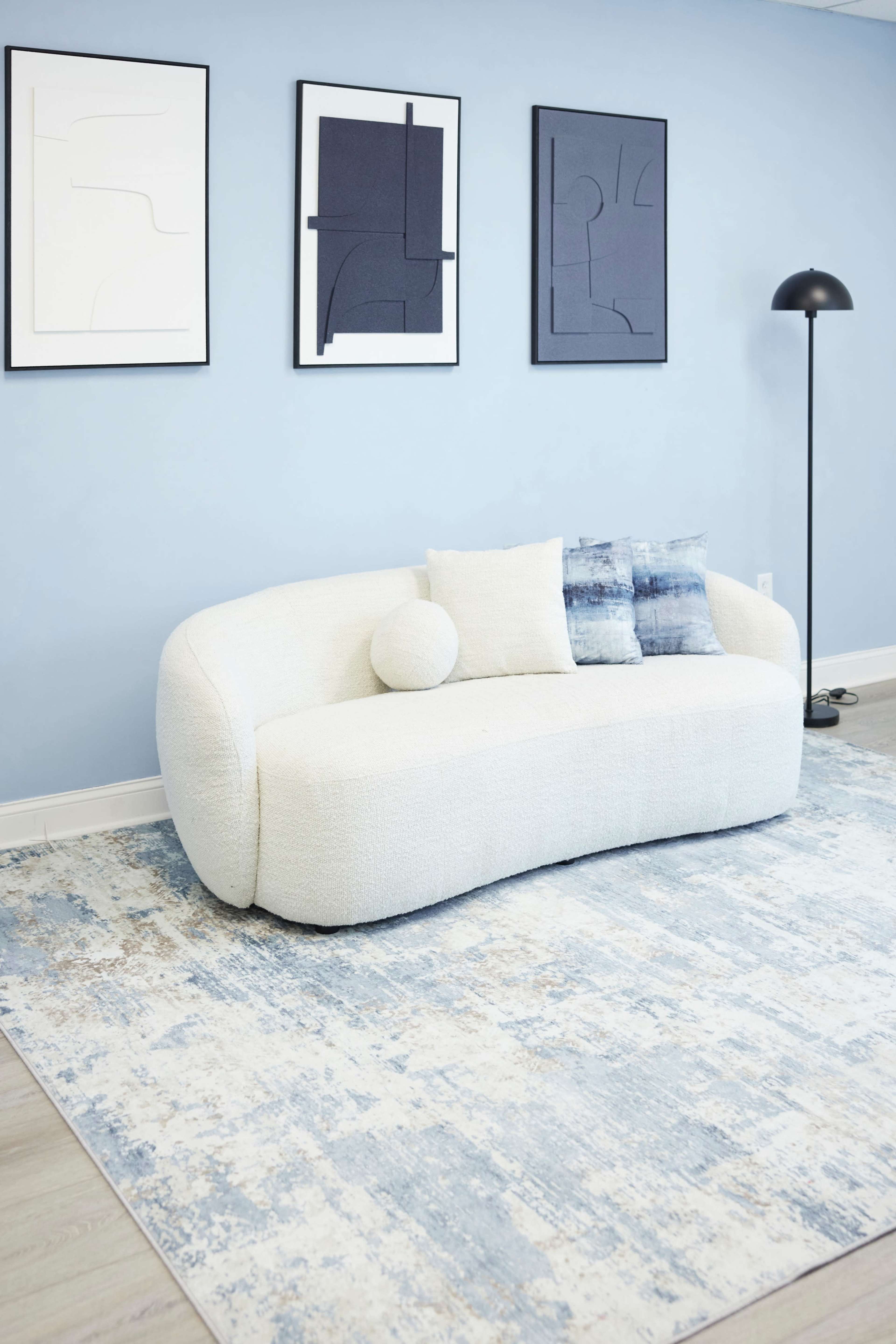 The image features a modern, curved white couch with a circular cushion and a small blue decorative pillow, positioned on a patterned blue area rug, against a light blue wall adorned with abstract art.