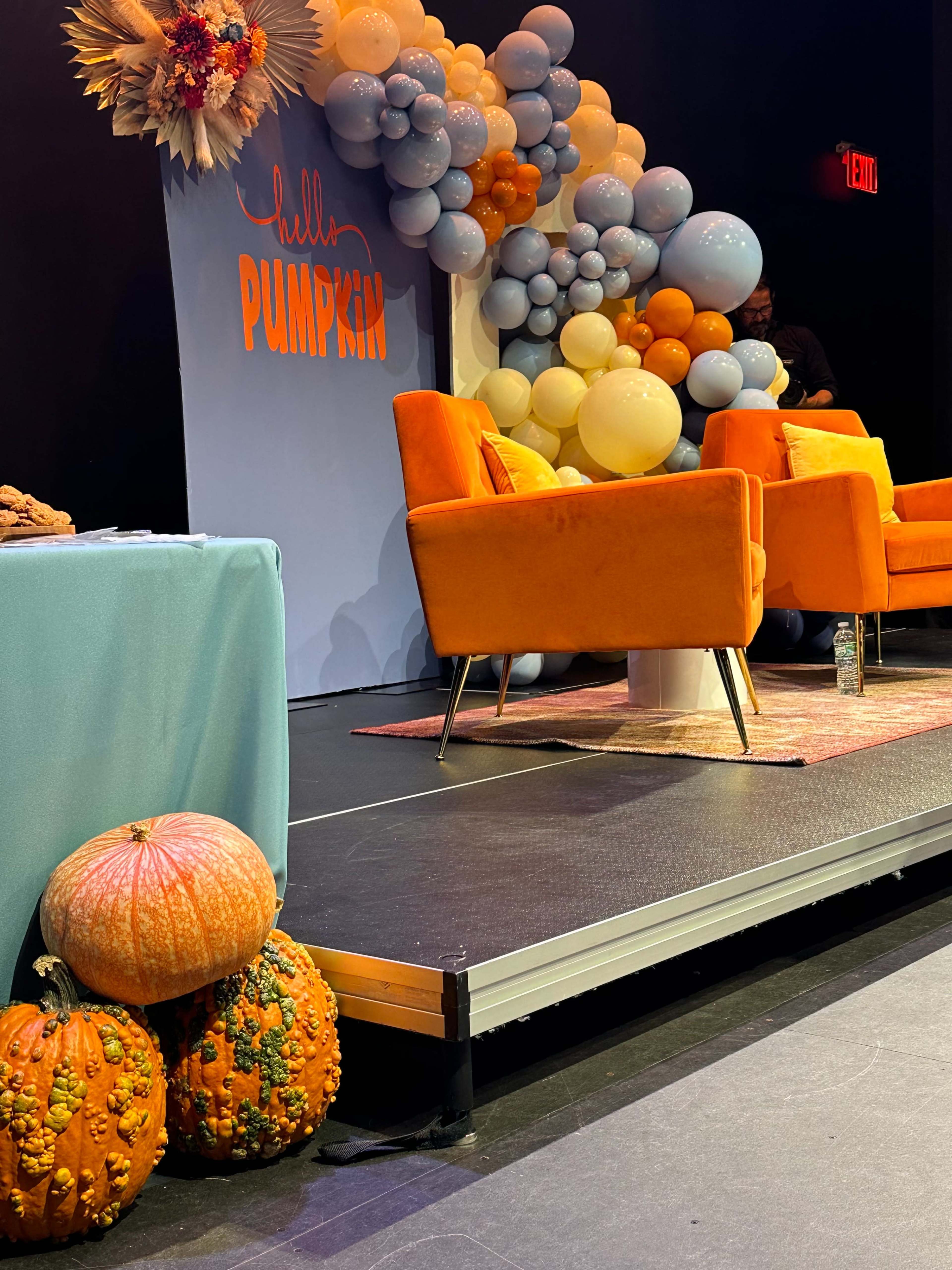 The image shows a stage setup with two orange chairs, a colorful balloon arrangement, and decorative pumpkins positioned nearby.