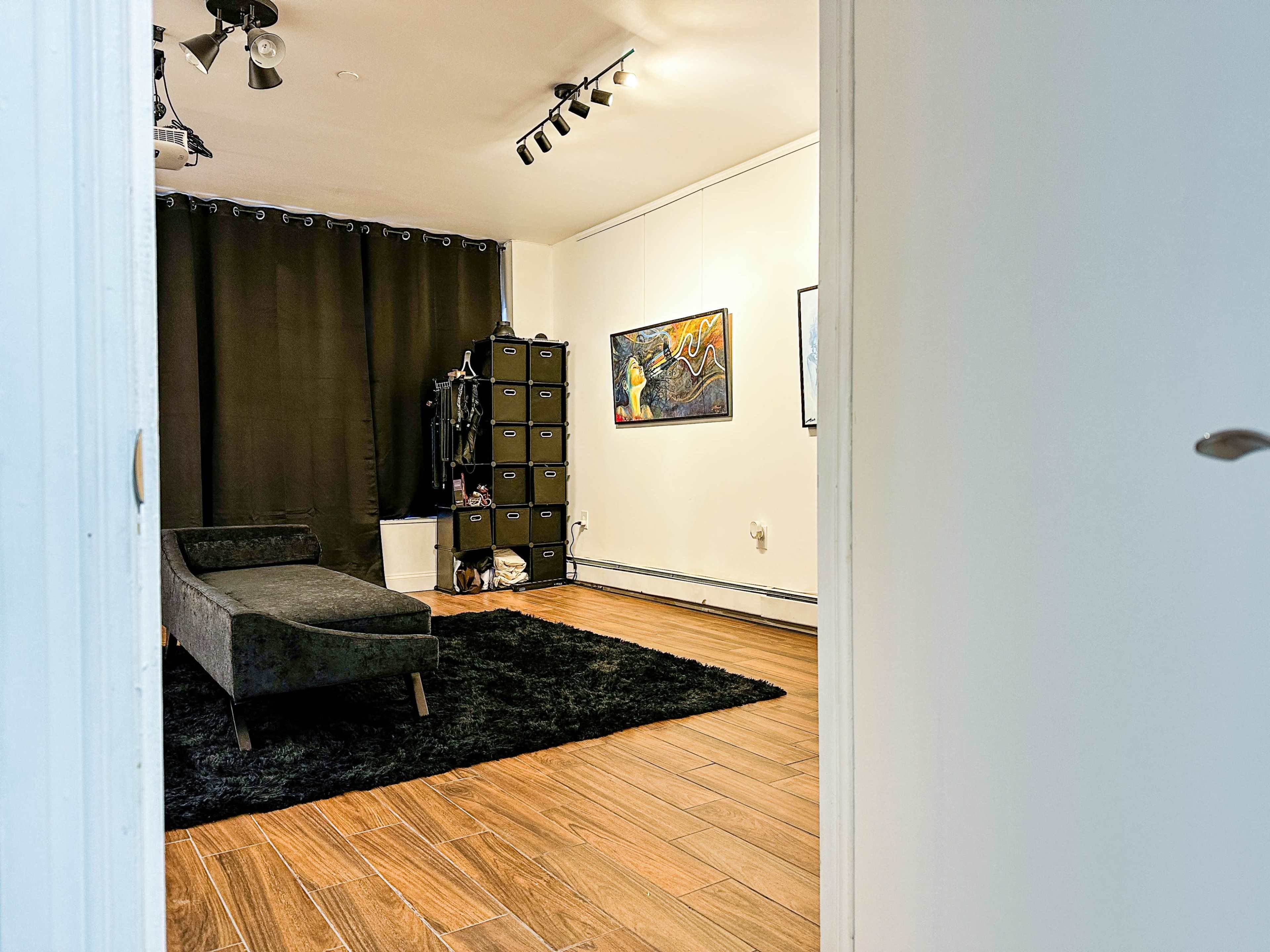 The image shows a spacious room with dark curtains, a black couch, a rug, storage boxes, and artwork on the walls.