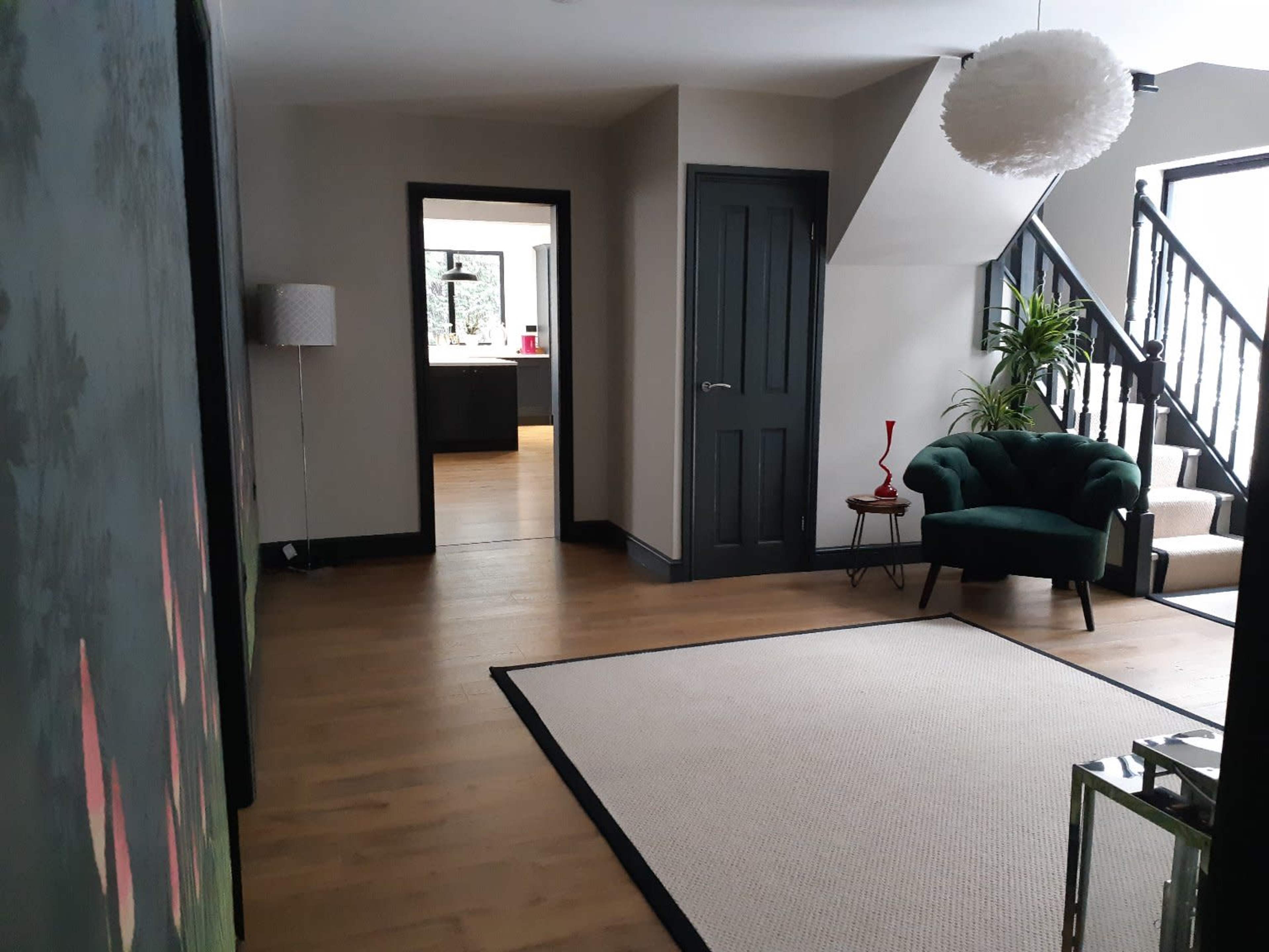 A modern hallway with wooden flooring, a green armchair, a staircase, and a doorway leading to a different room.