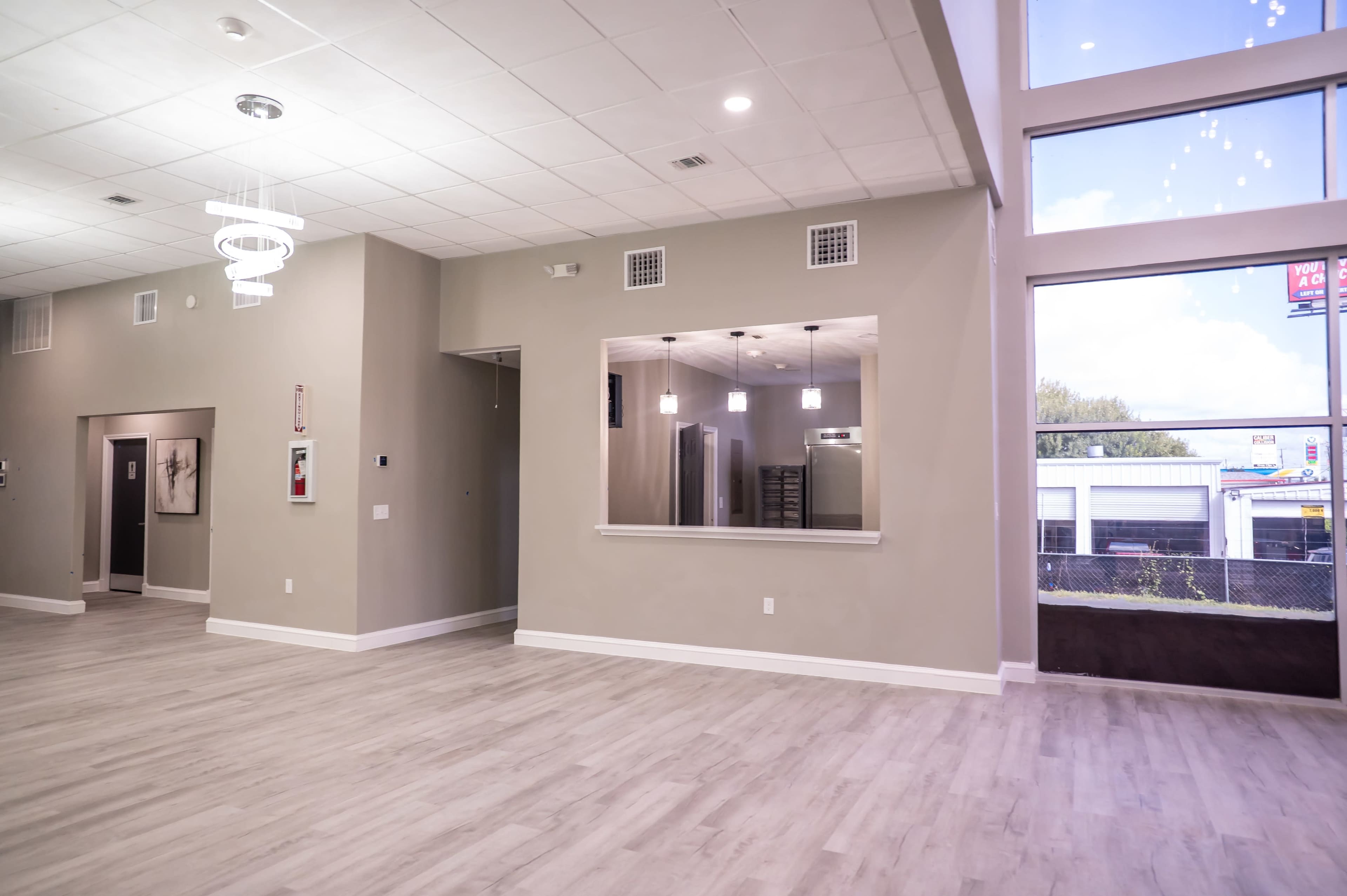 The image shows a spacious, well-lit interior room with light-colored walls and flooring, featuring large windows and a mirrored opening to another area.