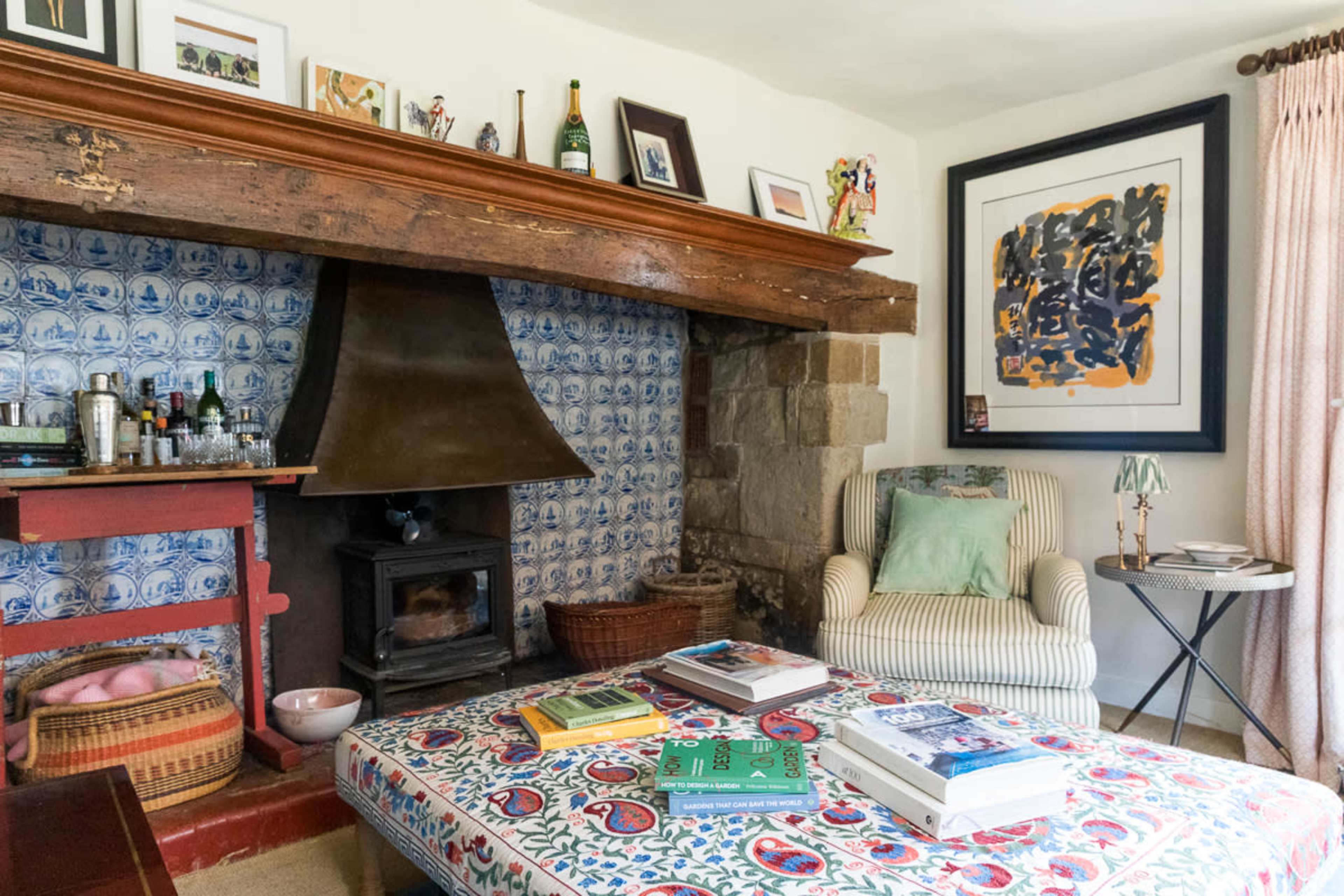 The image shows a cozy living room with a large wooden fireplace, patterned tile backdrop, and various books and items arranged on a coffee table.