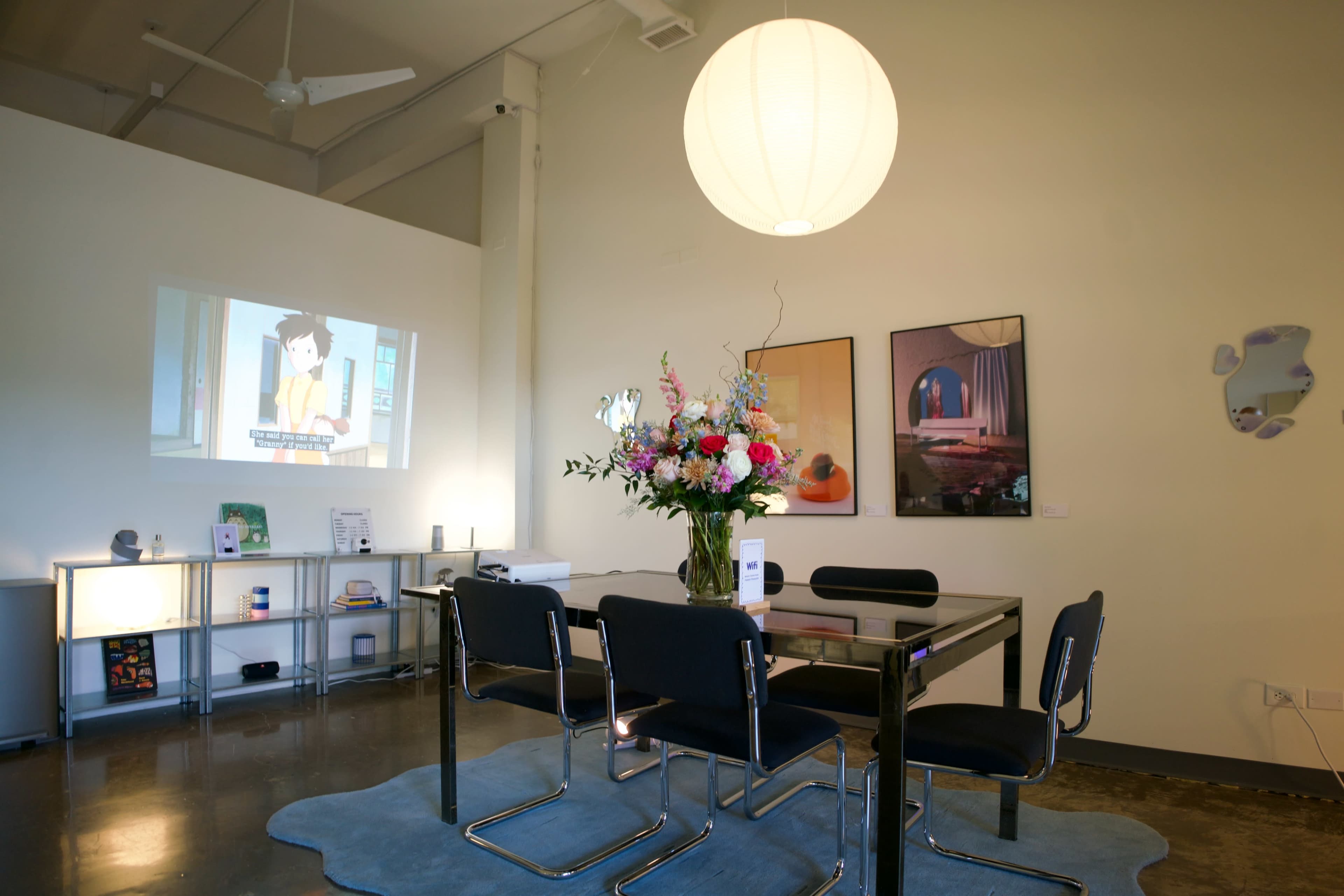 A modern meeting room features a glass table surrounded by black chairs, a colorful floral arrangement, and a projected screen displaying an animated scene.