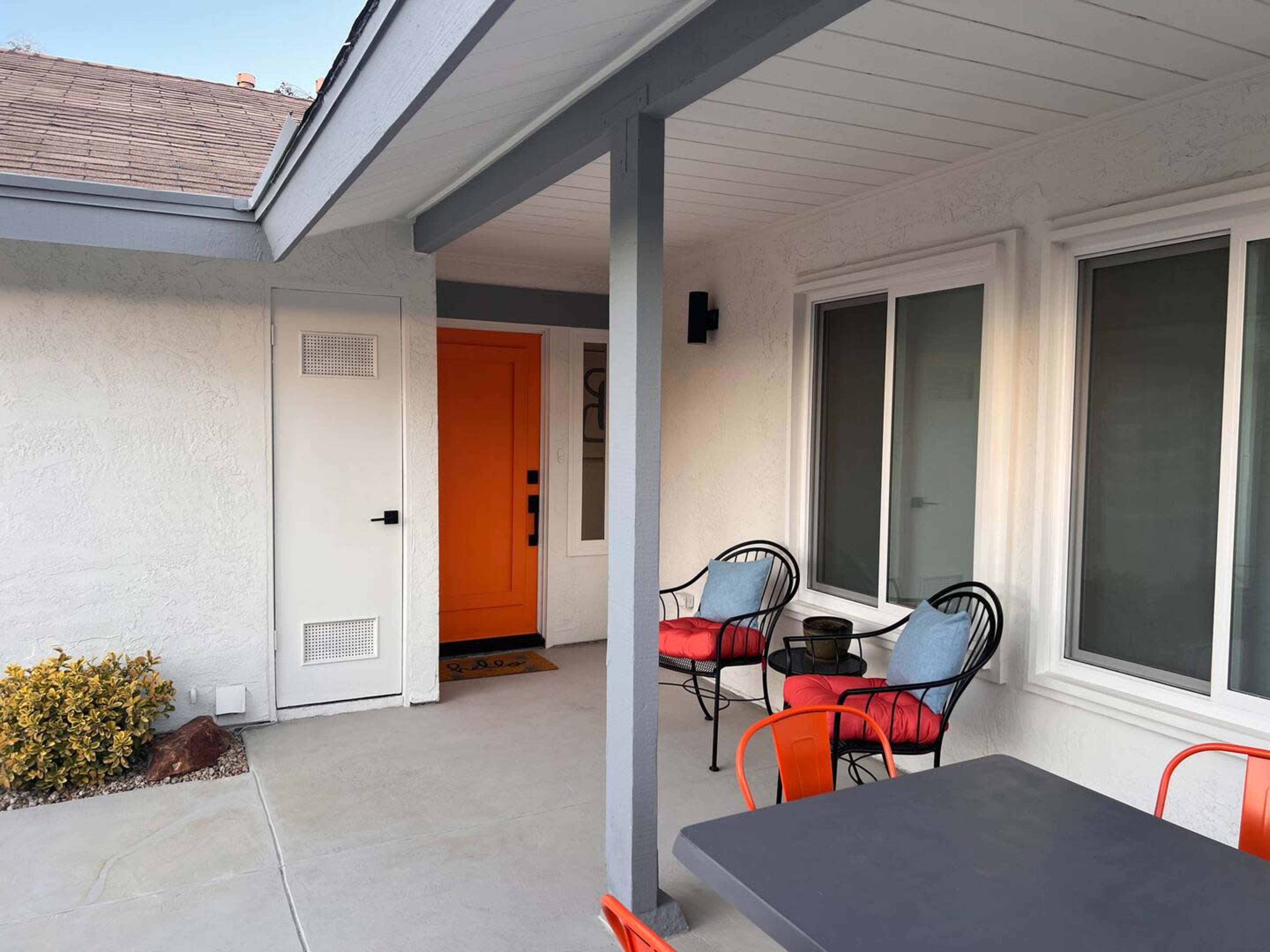 A porch area with a bright orange front door, two black chairs with cushions, and a table, adjacent to a wall with two windows.