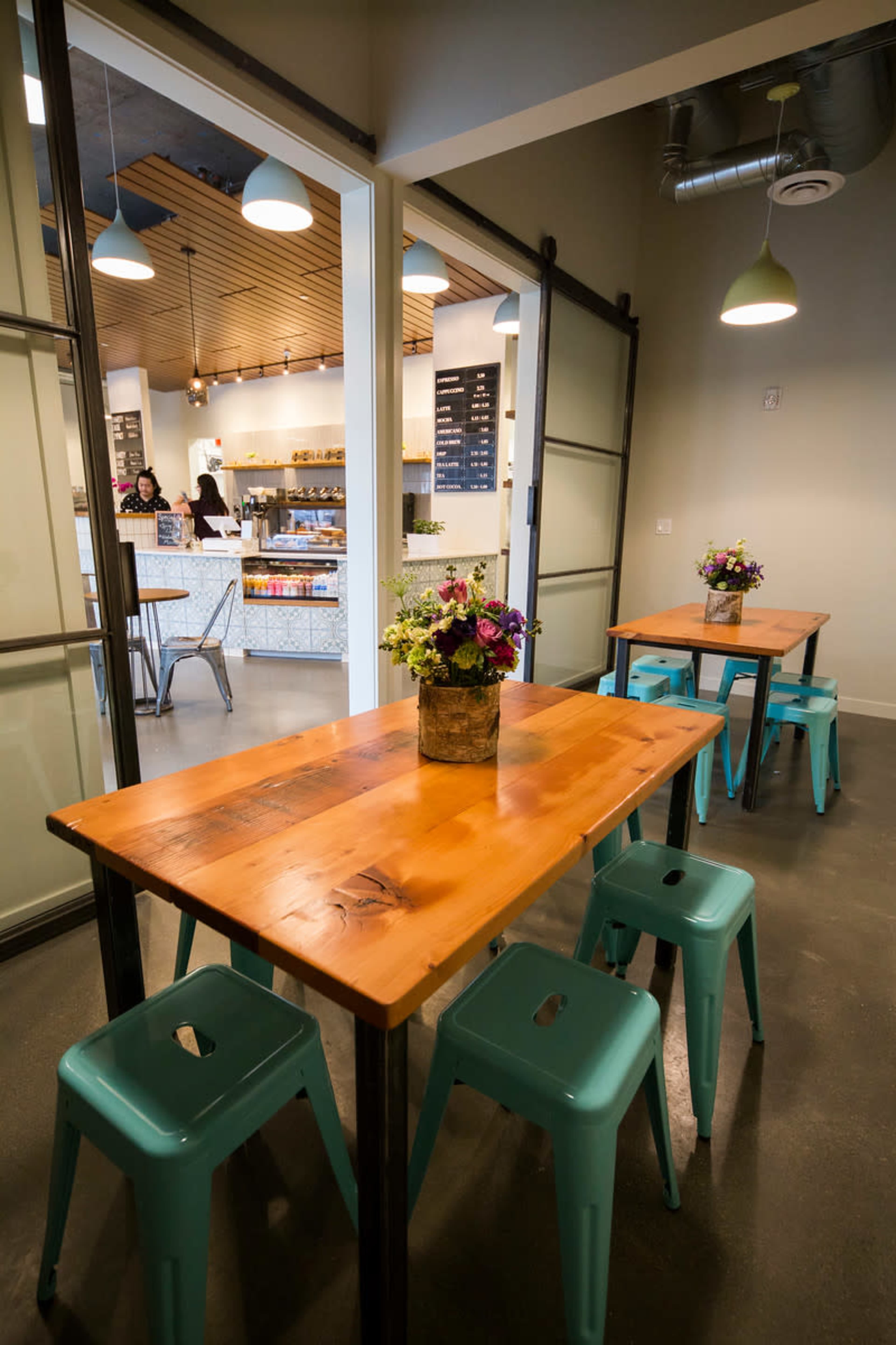 A modern cafe interior with wooden tables, turquoise stools, and a counter in the background where staff are preparing food.