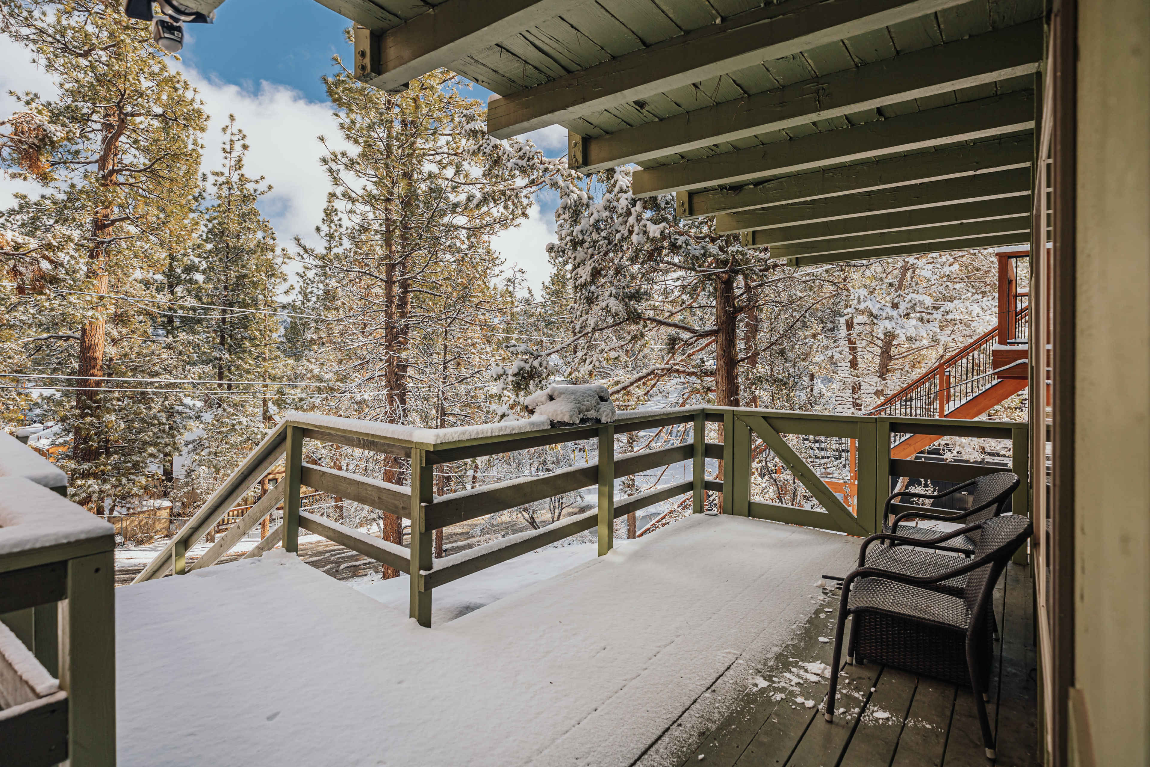 A snowy deck overlooks a forest of pine trees and a staircase leading to the ground below.