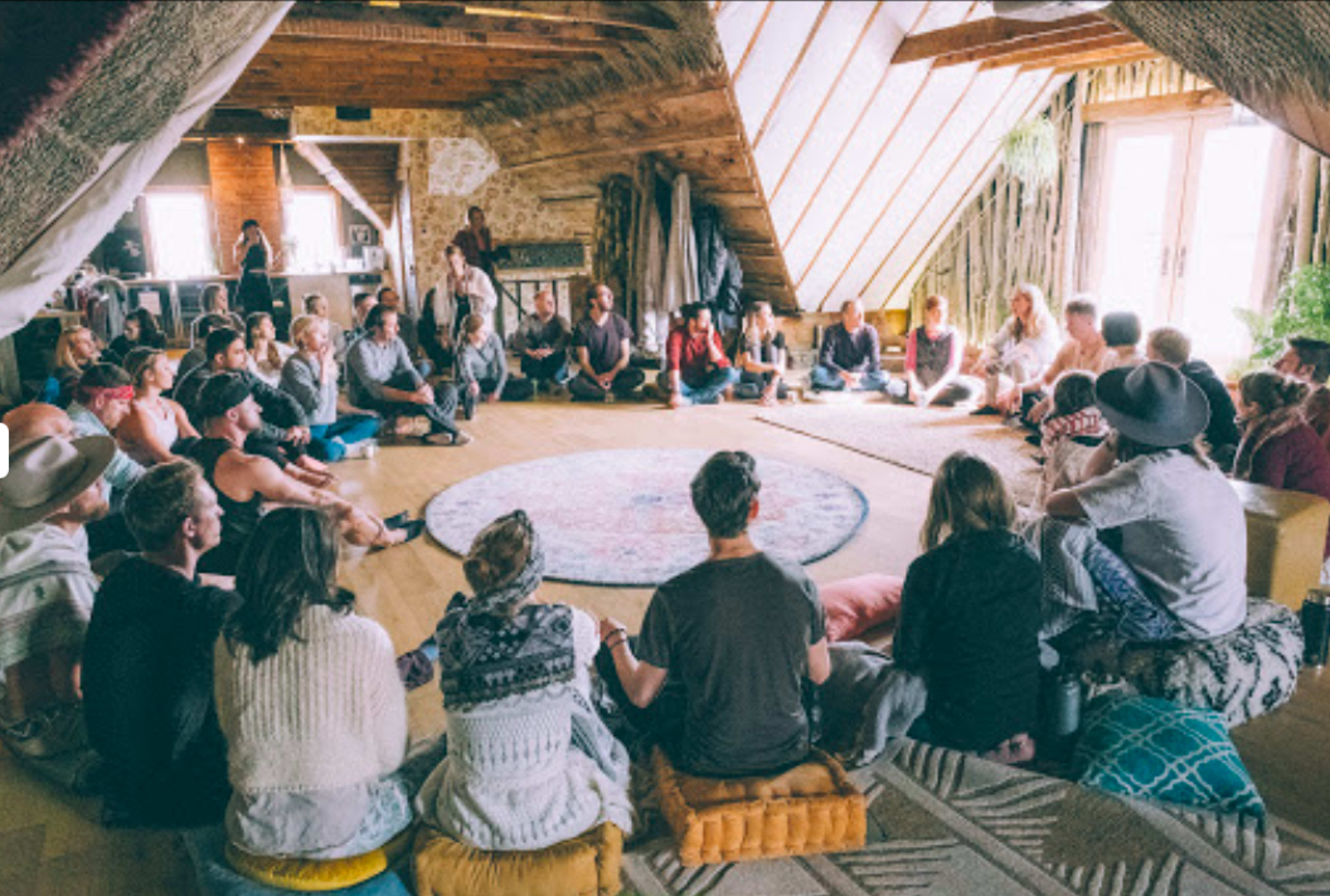 A large group of people sits in a circle on the floor of a rustic, well-lit room with a vaulted ceiling, engaged in a discussion.