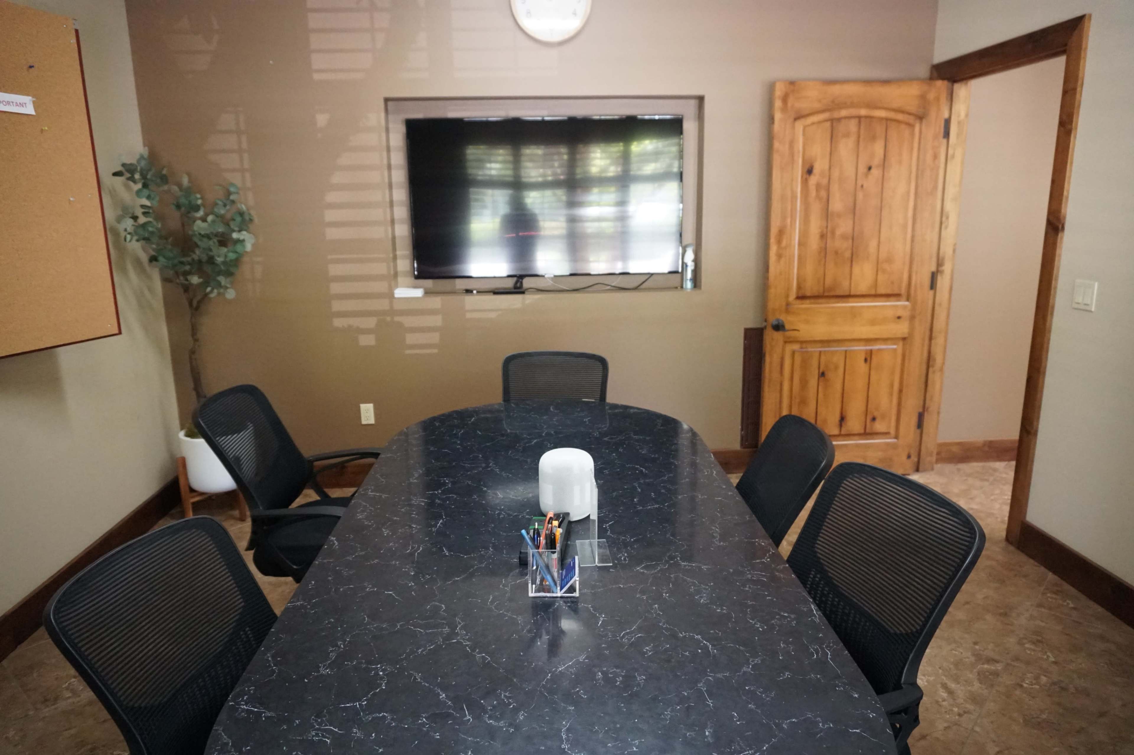 A conference room features a large dark table surrounded by black chairs, a wall-mounted TV, and an open door leading to another space.