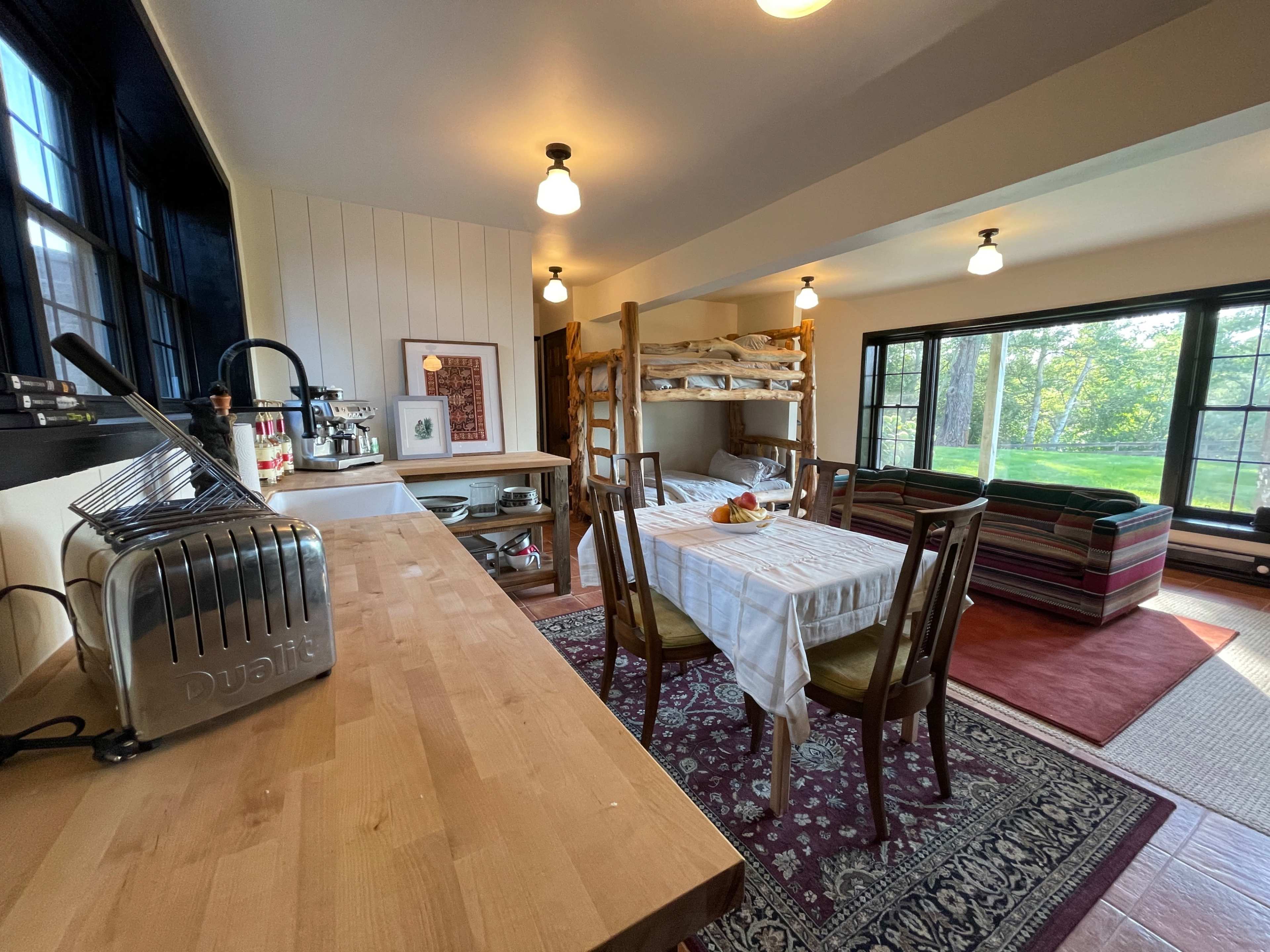 A cozy, open-concept living space featuring a kitchen area with a wooden table, a small dining area, and a bunk bed beside large windows that provide a view of the outdoors.