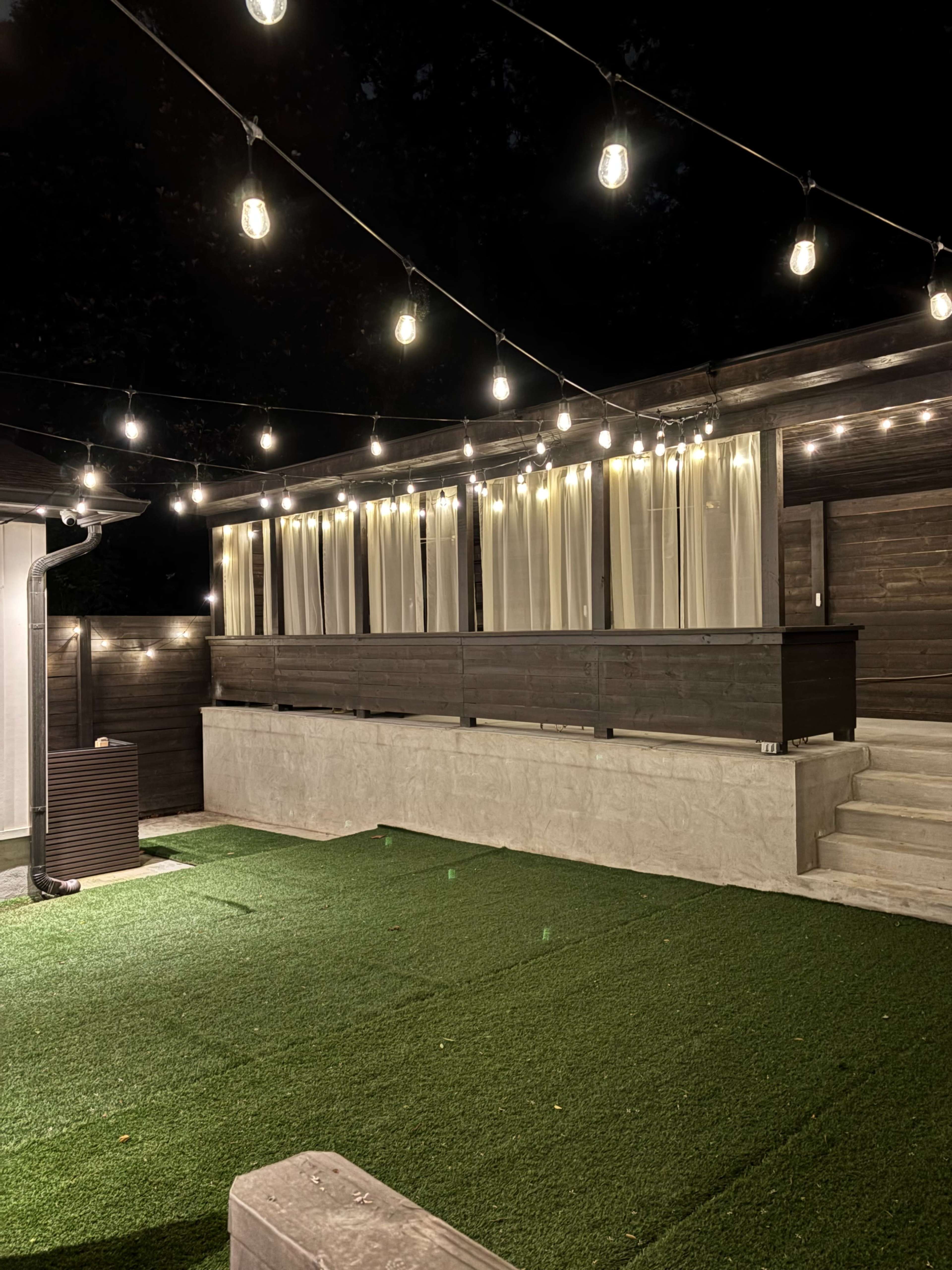 The image shows a backyard patio at night, illuminated by string lights, with a rectangular area of artificial grass and a wooden deck in the background.
