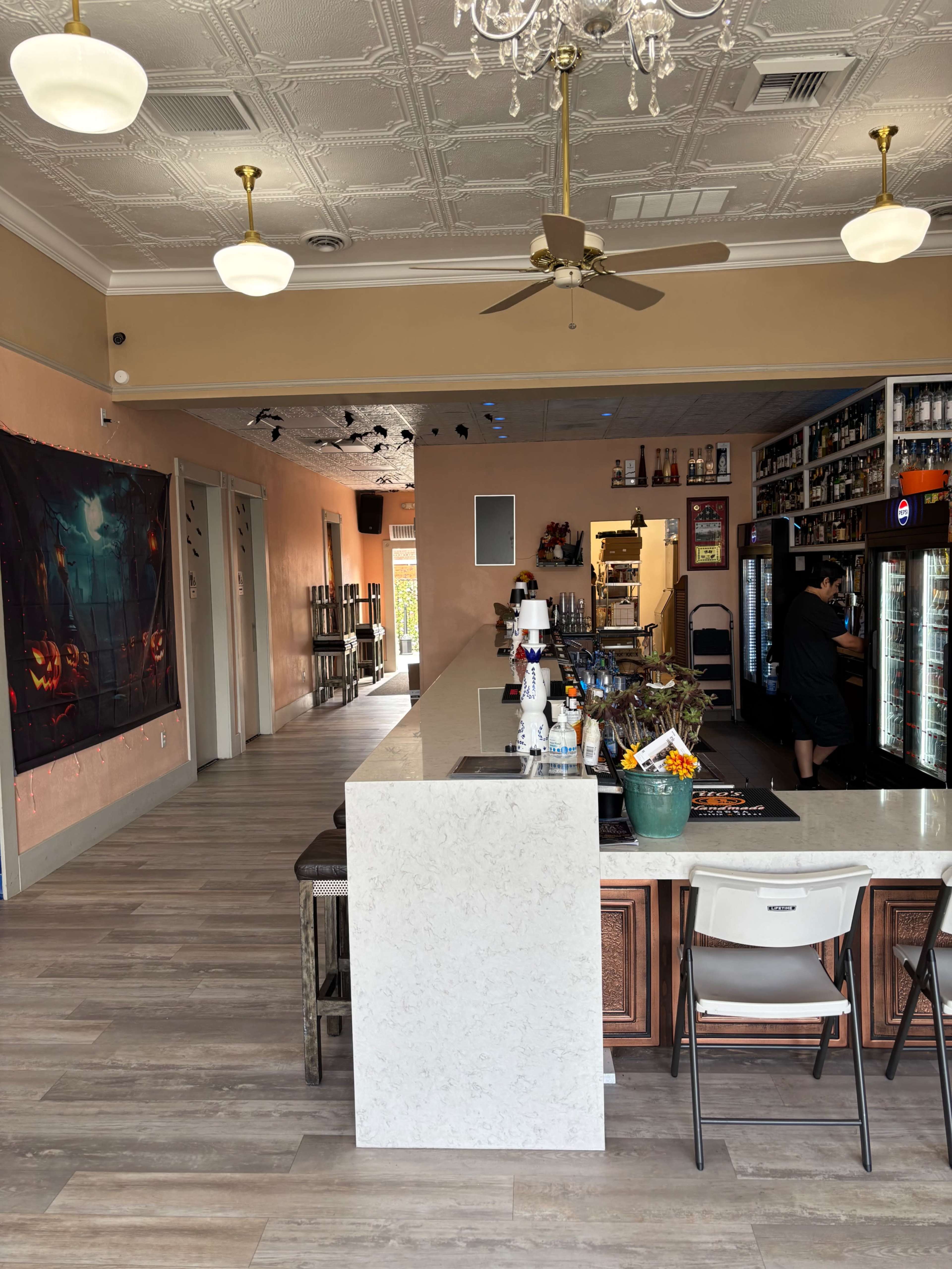The image shows a partially filled bar area in a café, with a counter on one side, a beverage cooler, and shelves stocked with bottles in the background.