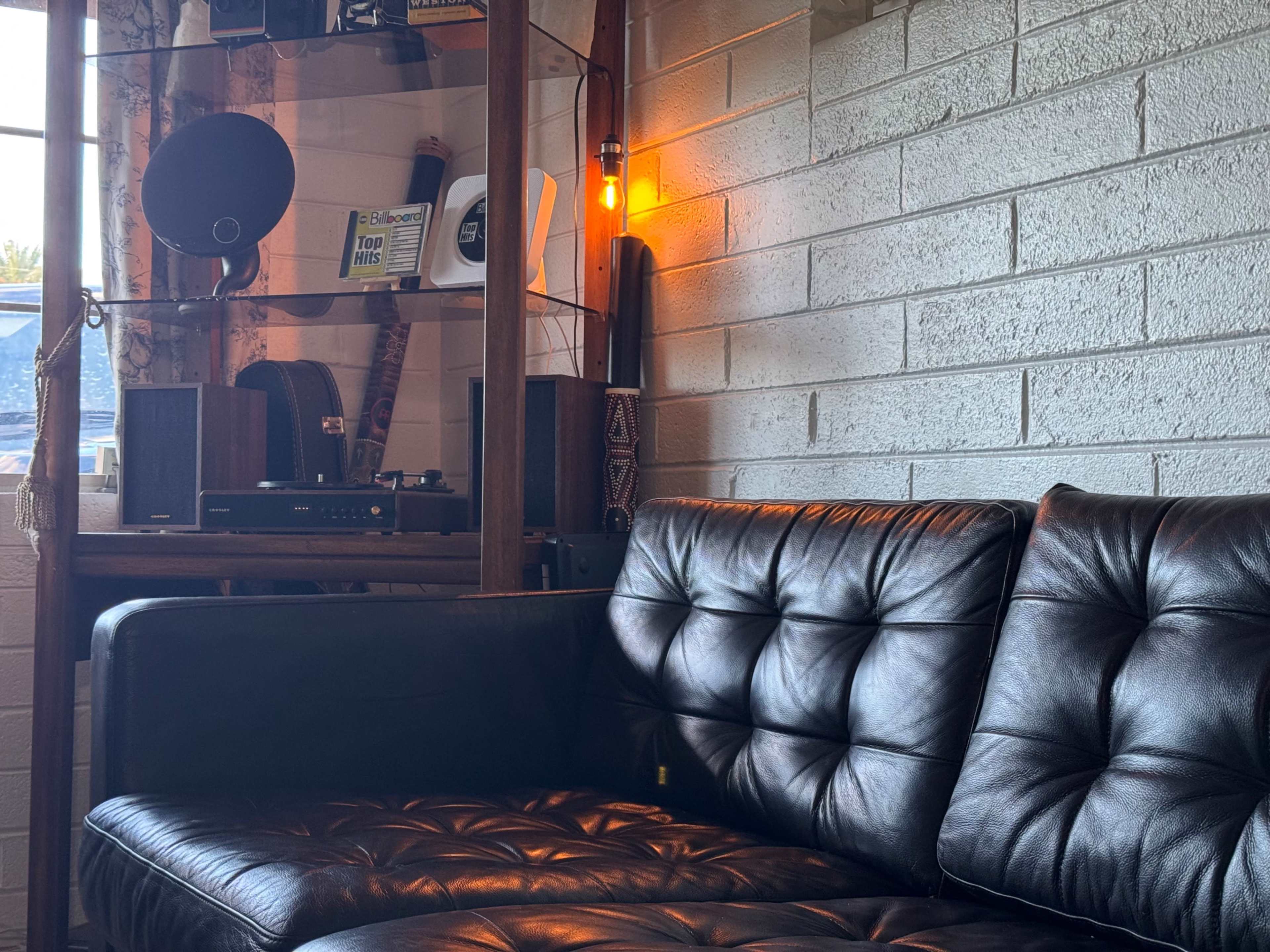 The image shows a leather sofa positioned beside a wall, with a wooden shelf featuring various audio equipment and a warm light source illuminating the space.