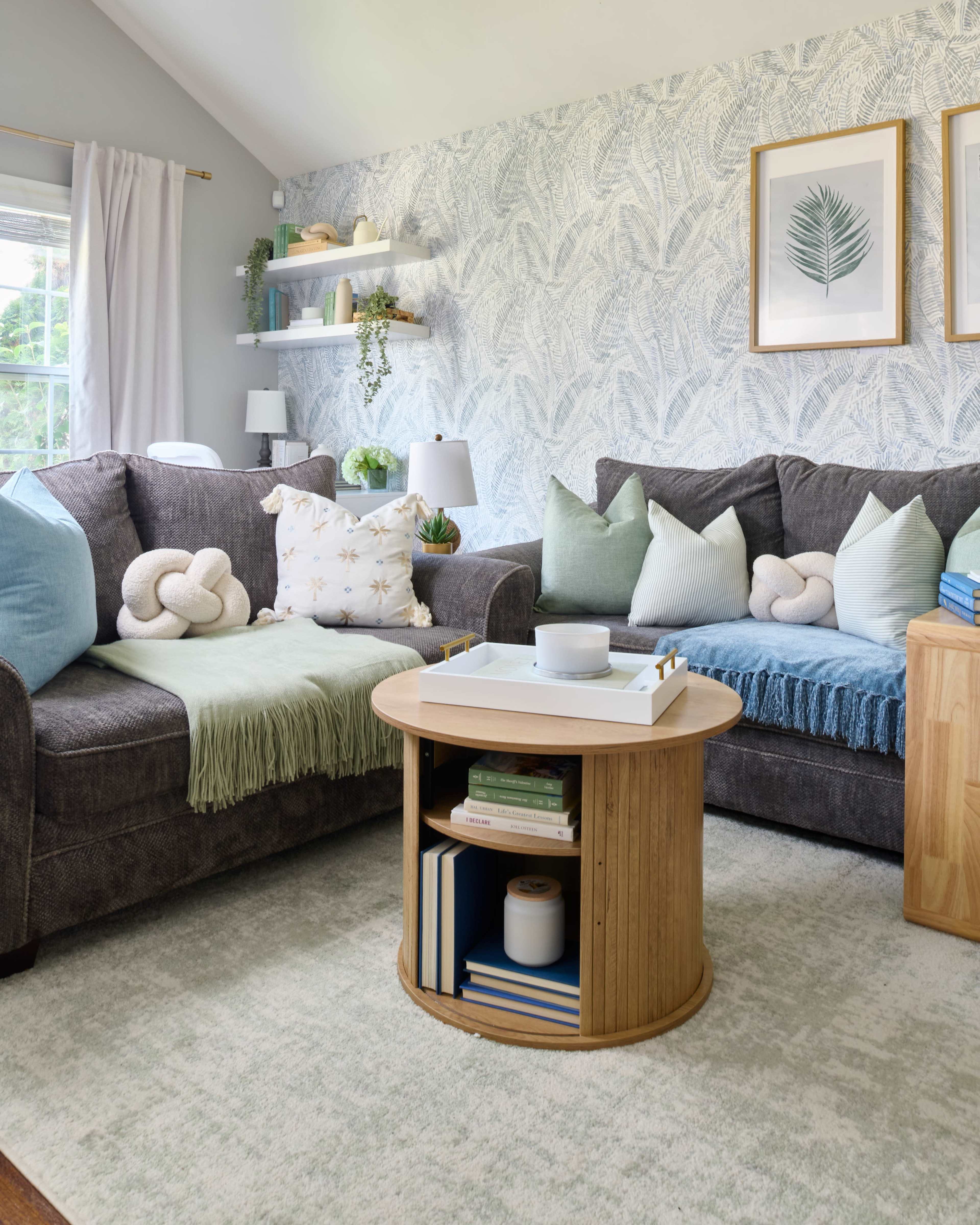 The image shows a cozy living room featuring two dark gray sofas adorned with various cushions, a round wooden coffee table with books and a decorative item, and light-colored walls decorated with patterned wallpaper and framed artwork.