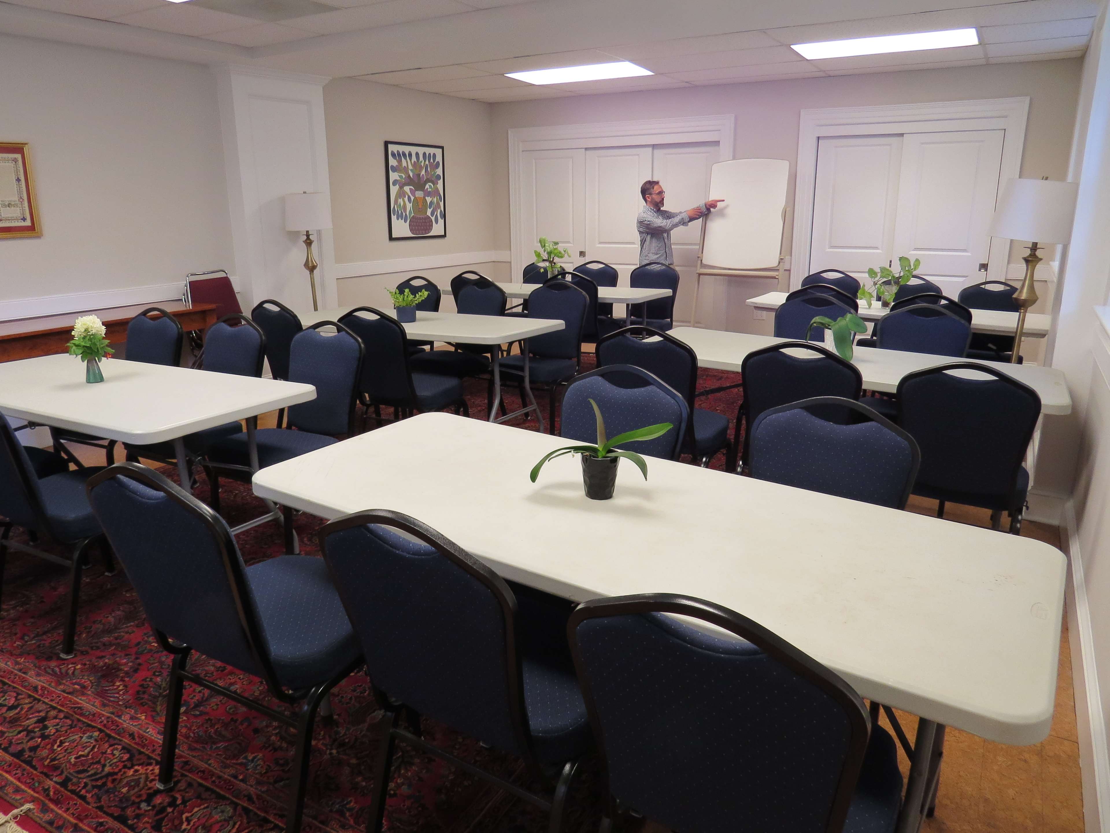 A person is setting up a meeting room with tables and chairs arranged around a central area, framed by decorative lamps and plants.