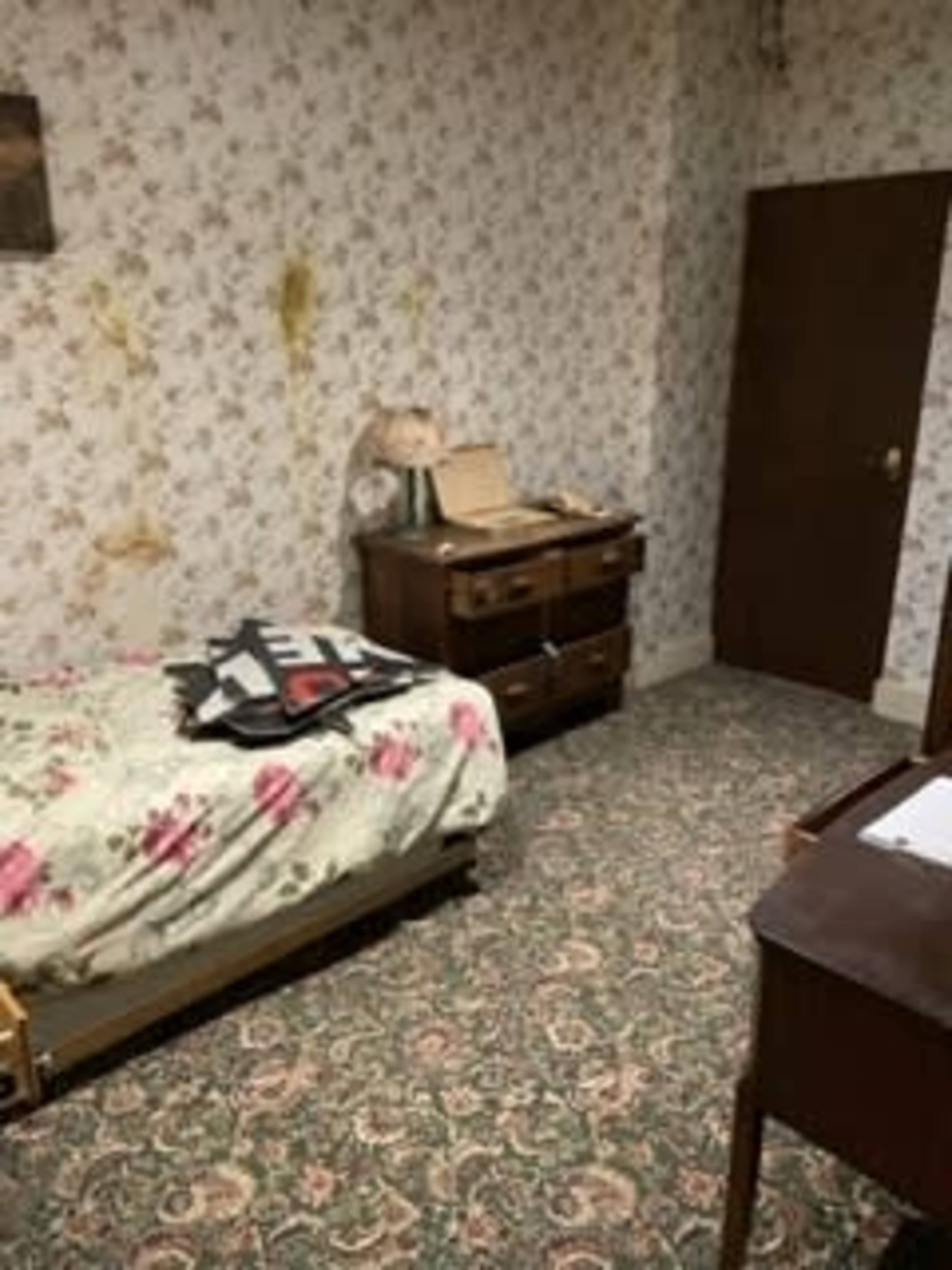 The room features a floral-patterned bedspread on a bed, a wooden dresser, a lamp, and a patterned carpet against wallpaper adorned with a floral design.