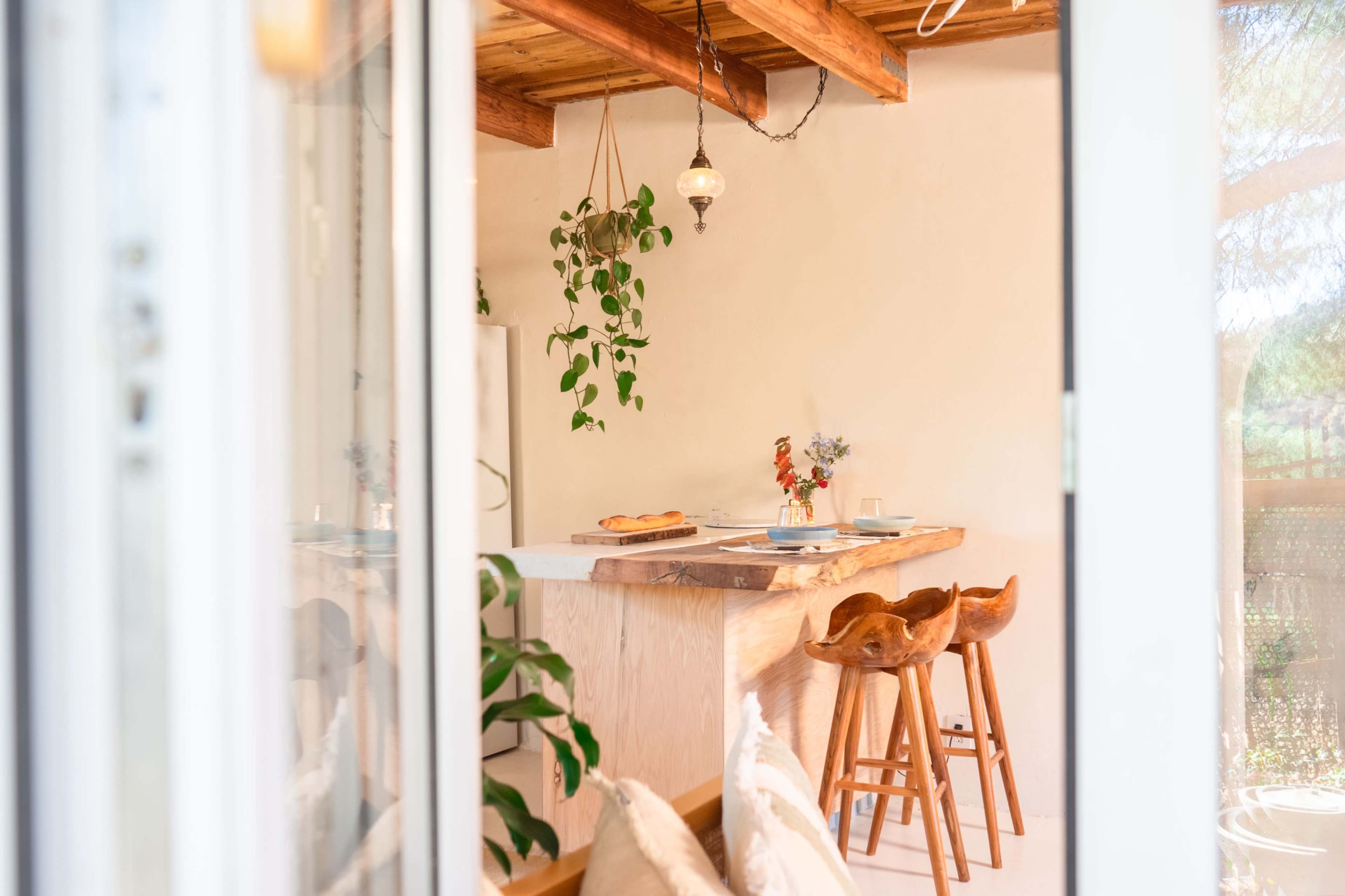 A bright kitchen features a wooden island with bar stools, potted plants, and a dining setup visible through a glass door.