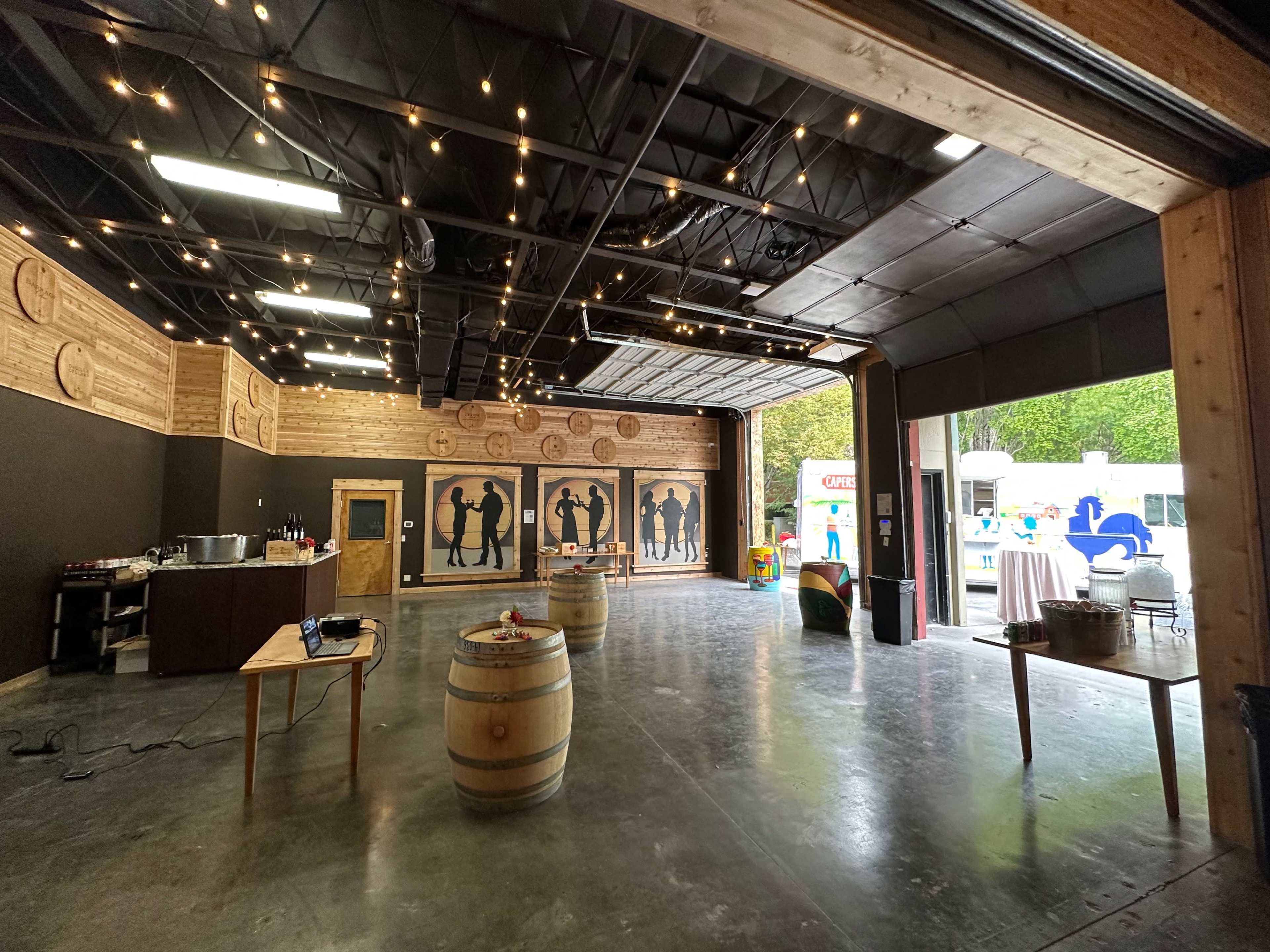An indoor event space features wooden walls, silhouetted figures, wine barrels, and string lights.