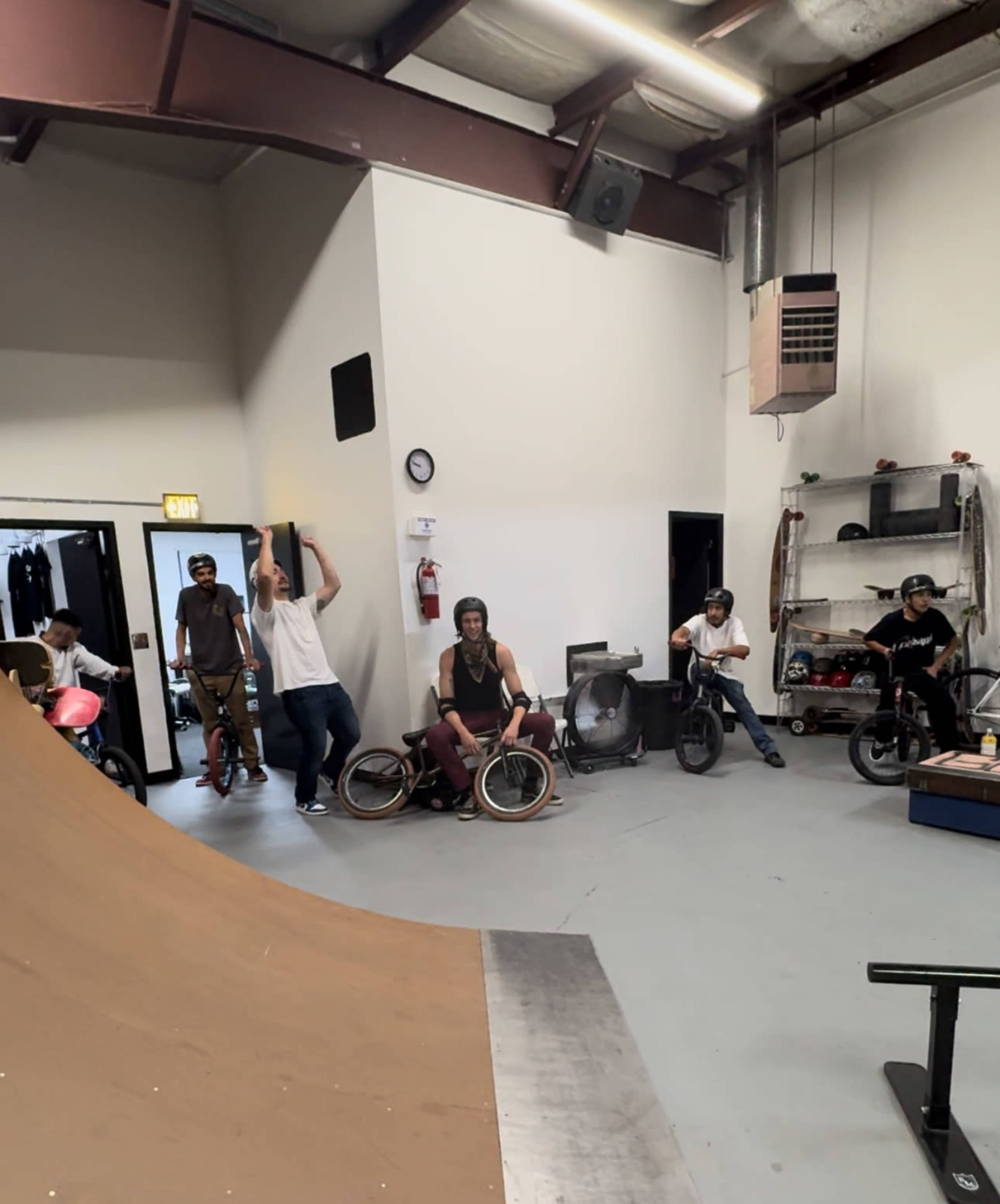 A group of five people is gathered inside a skatepark area, some on bicycles and others standing near a half-pipe ramp.