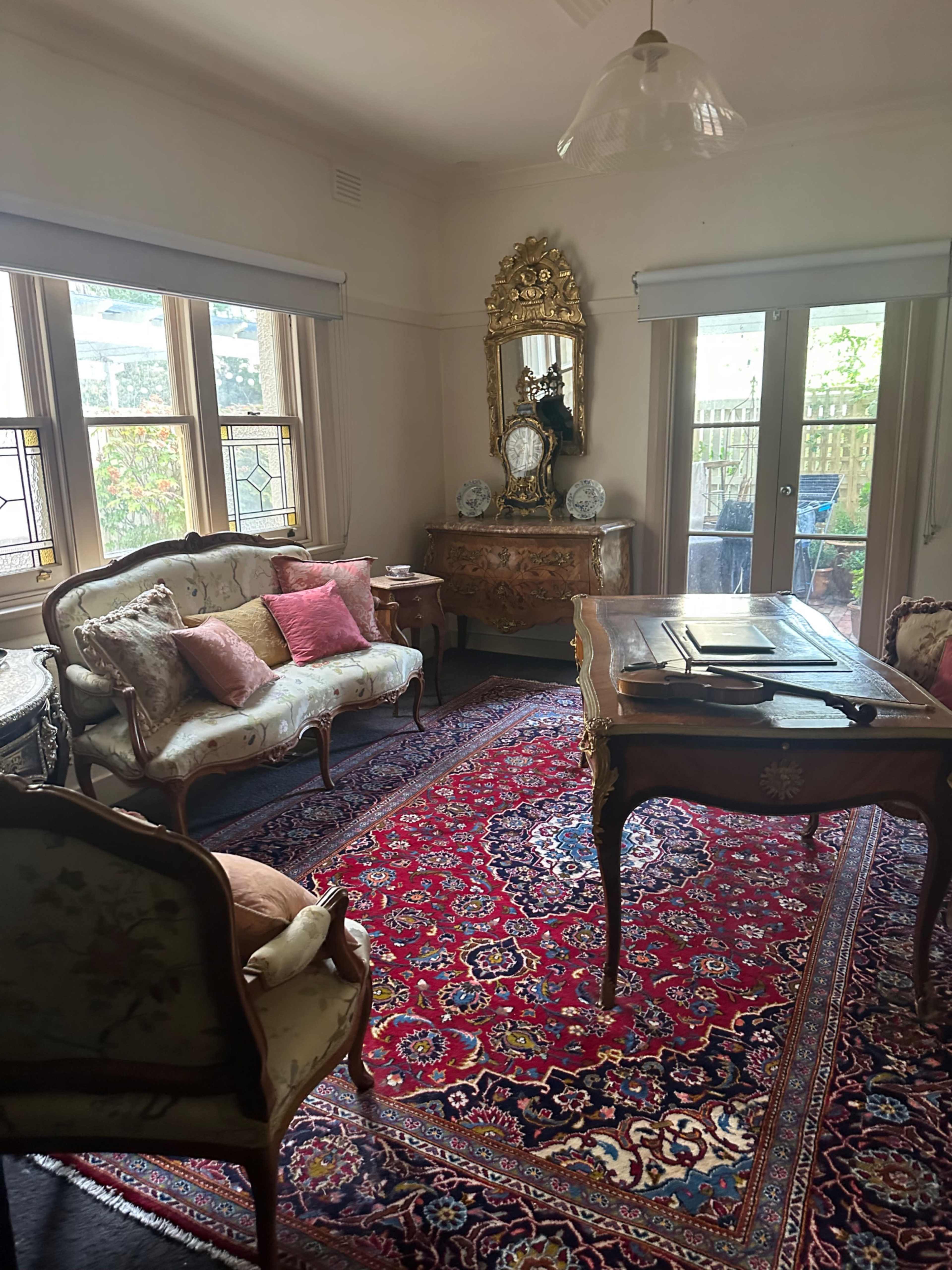 The room features a vintage sofa and armchair, an ornate mirror on a decorative dresser, and a large area rug under a wooden desk.