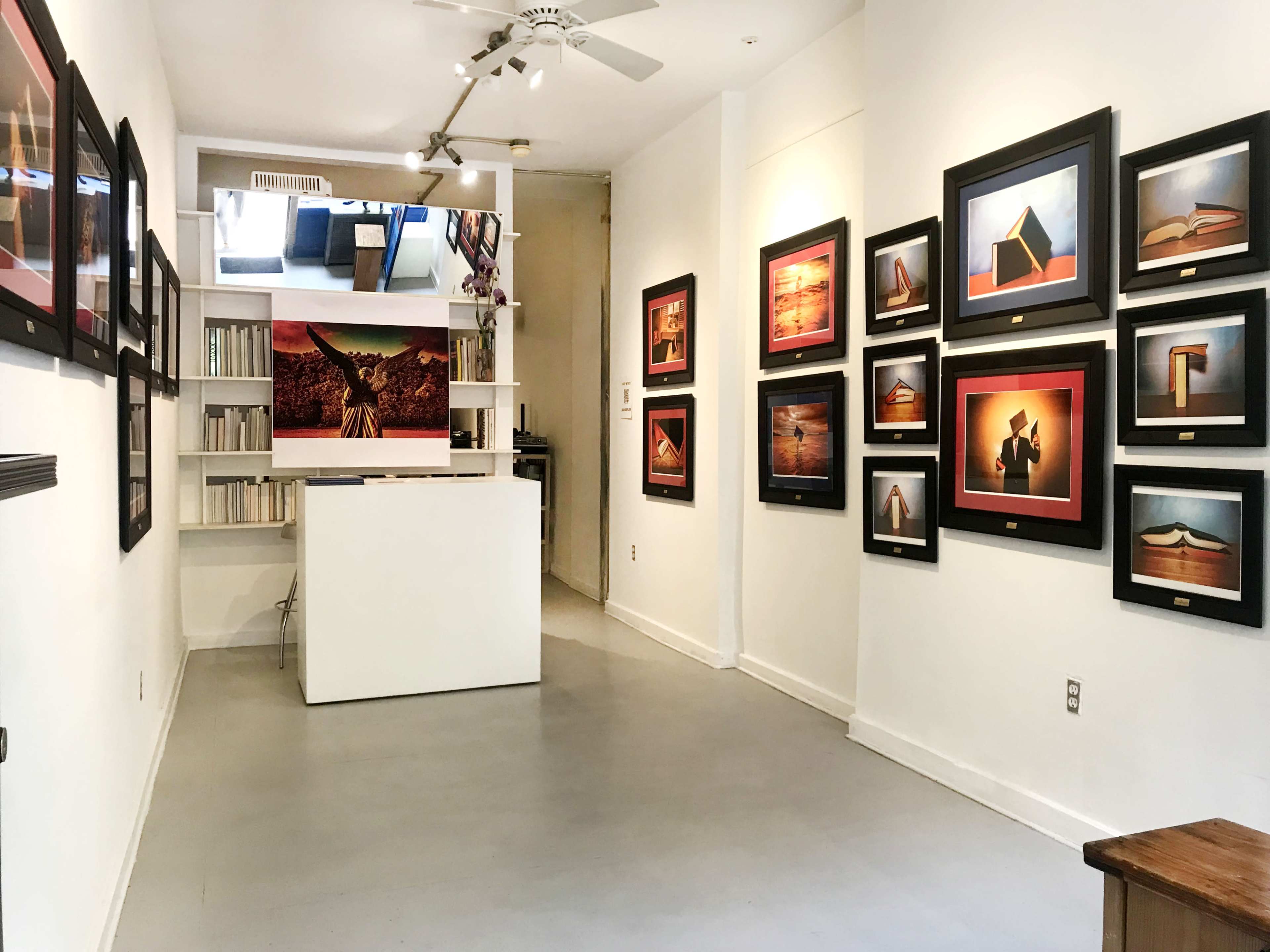 The image shows a gallery interior with framed artwork displayed on the walls, a white counter in the center, and shelves of books in the background.