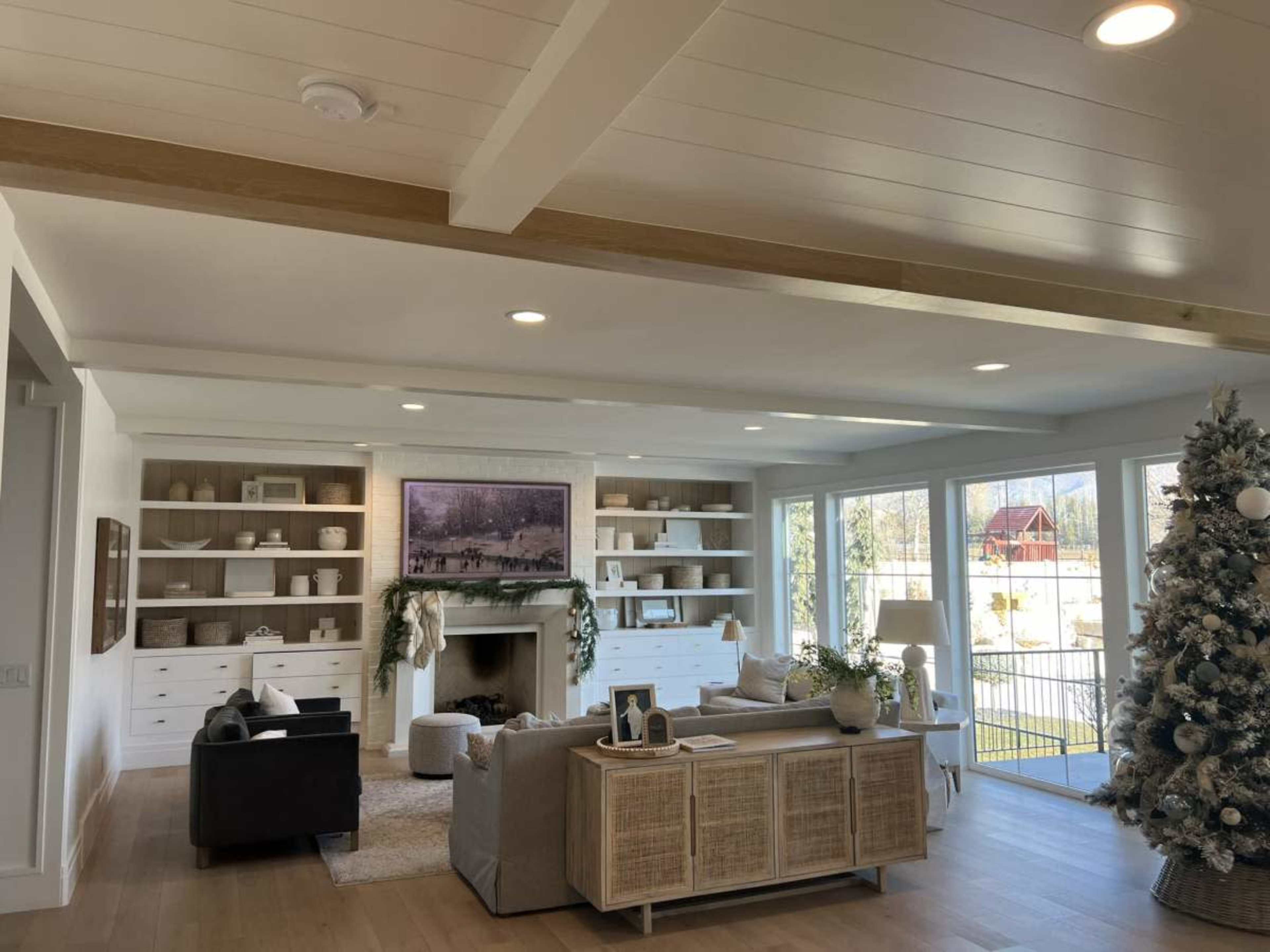A bright living room with a fireplace, built-in shelves, a Christmas tree, and large windows allowing natural light to fill the space.