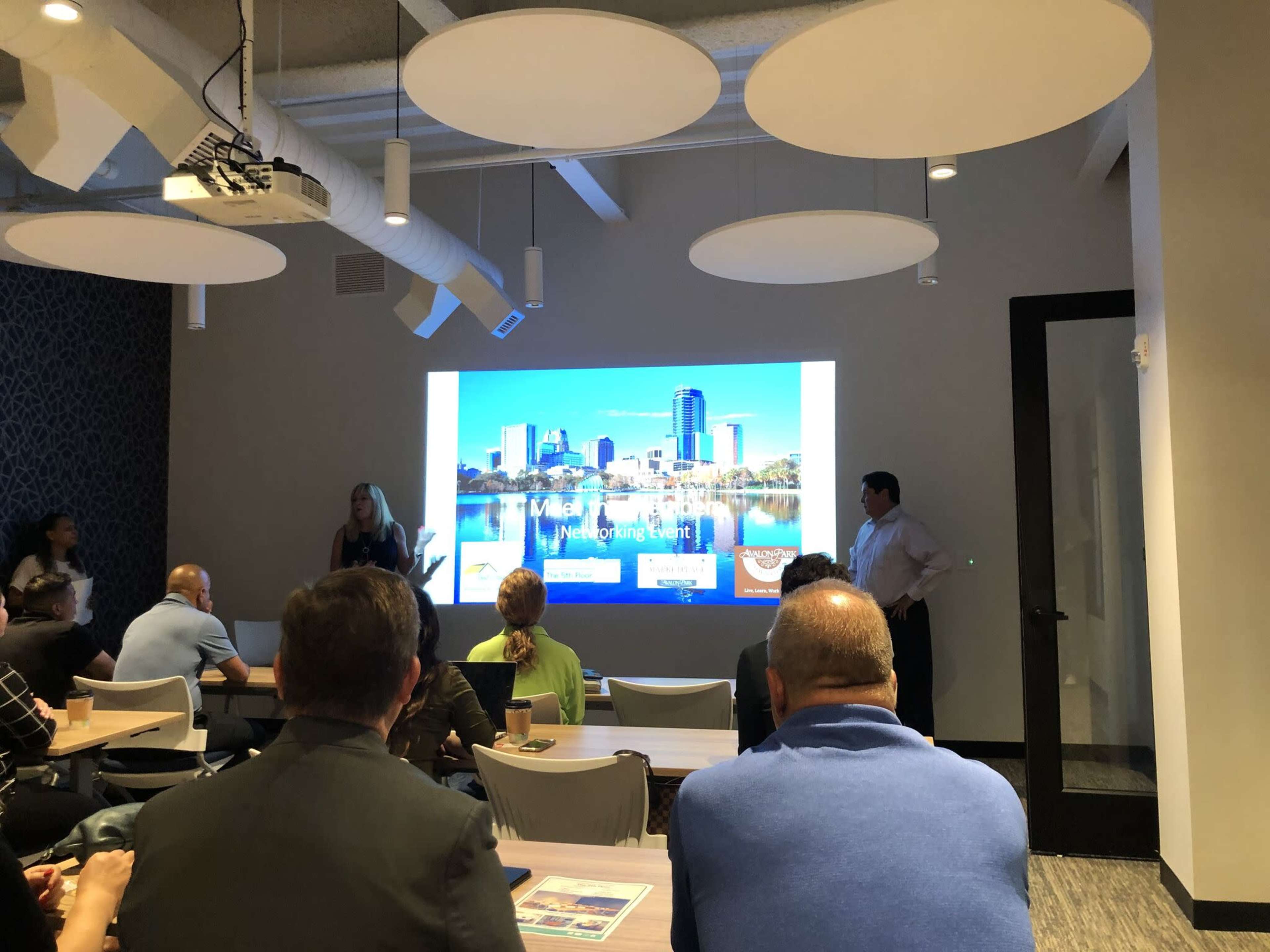 A group of people sits in a conference room, watching a presentation on a large screen that displays a city skyline.