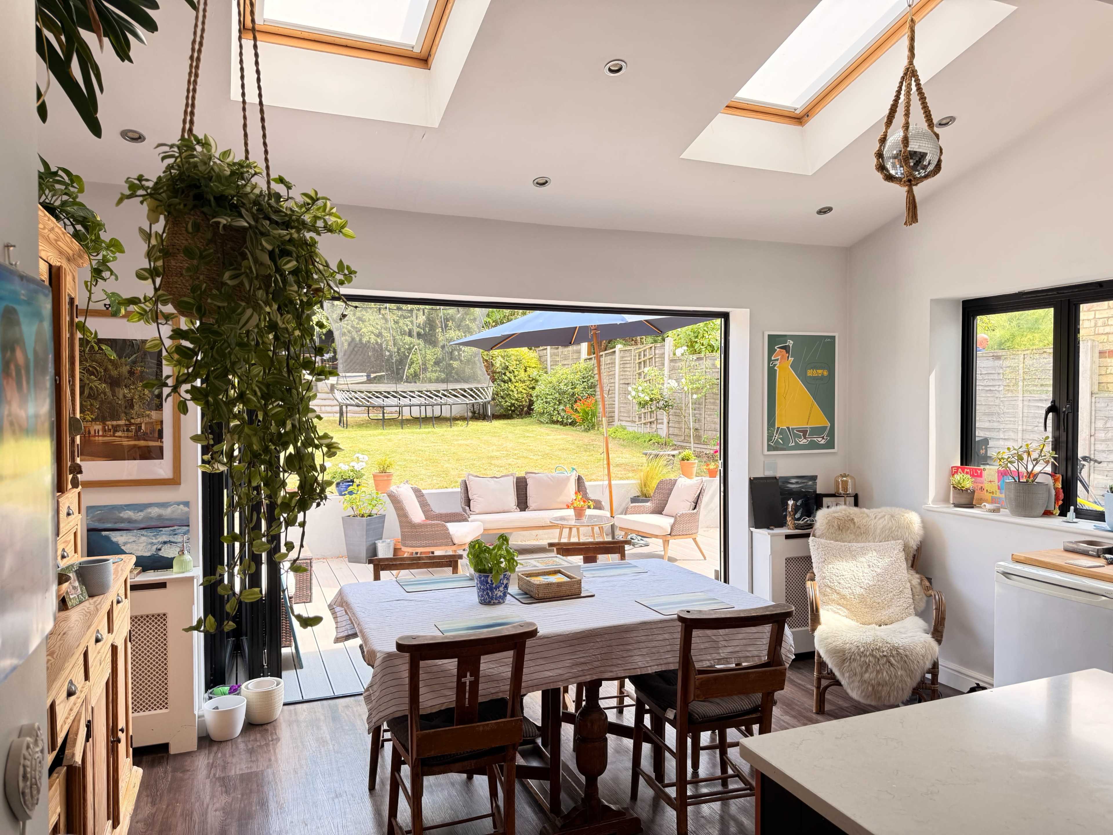 The image shows a modern kitchen with a dining table, bright natural light streaming in through skylights, and an open view of a grassy backyard with seating areas.