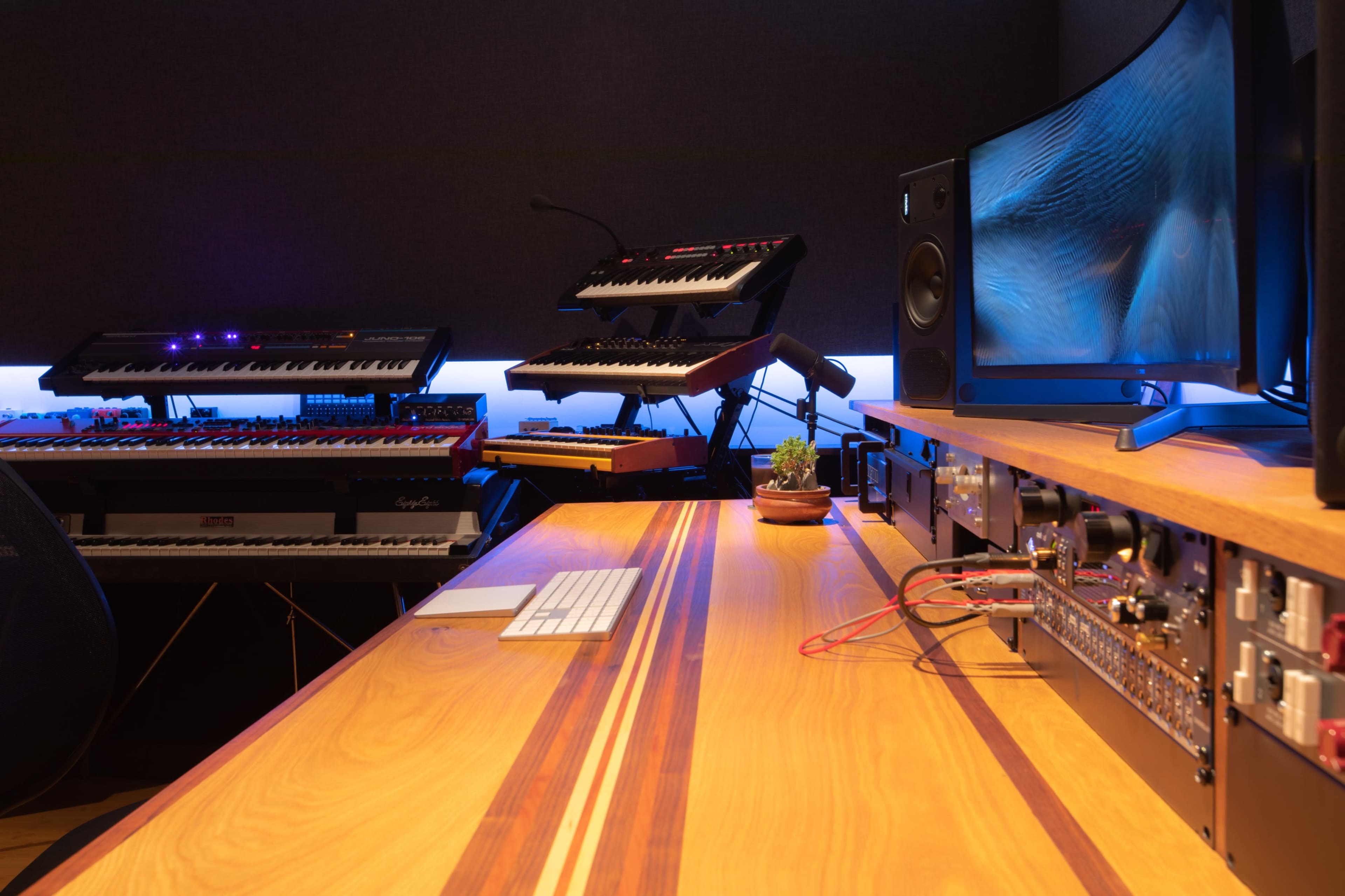 A modern music production studio featuring multiple synthesizers, a computer monitor, and a clean wooden desk with a keyboard and audio equipment.
