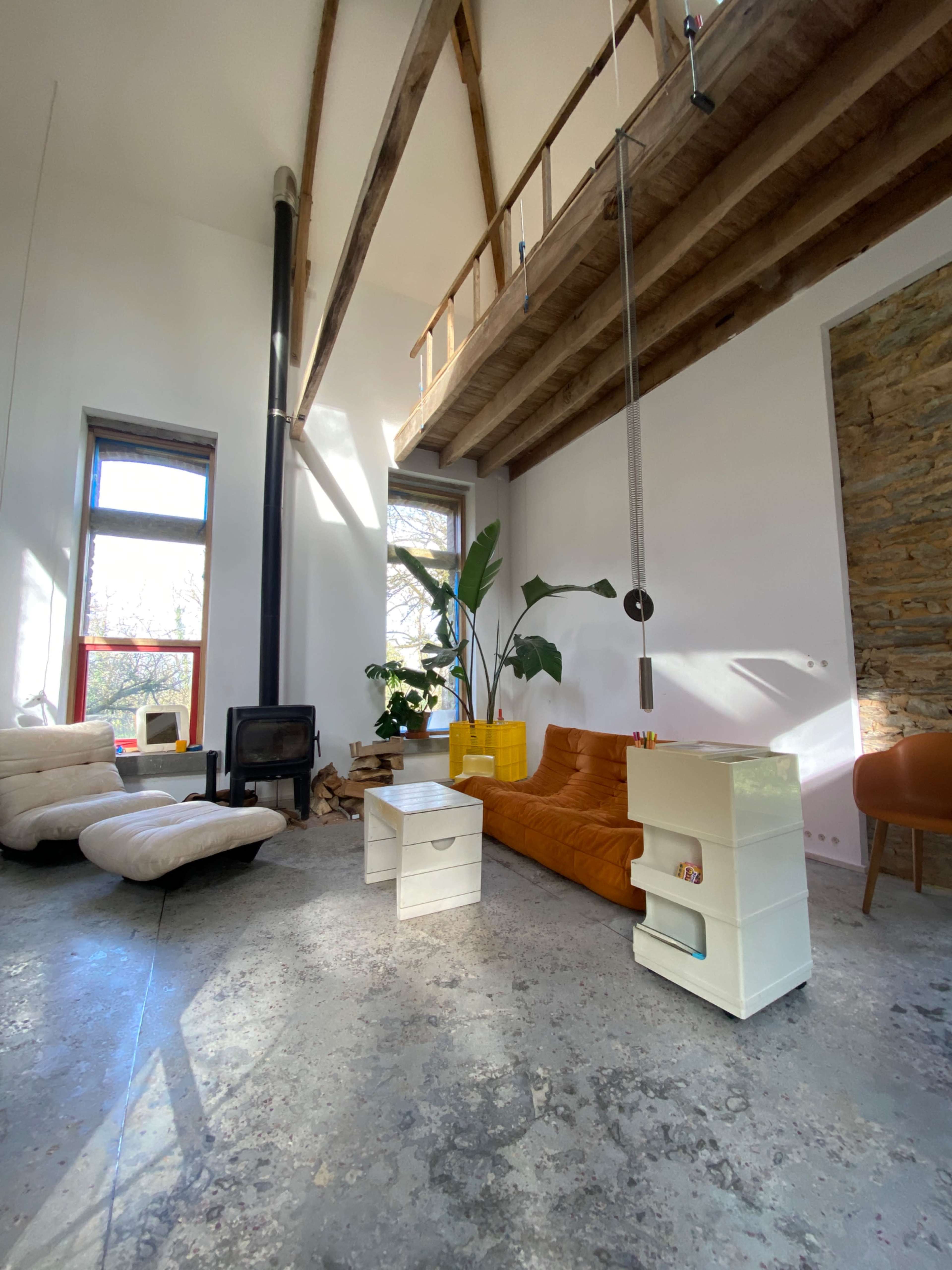 The image shows a spacious, modern living room with a brown sofa, a light-colored armchair, a small white table, and large windows that let in natural light.