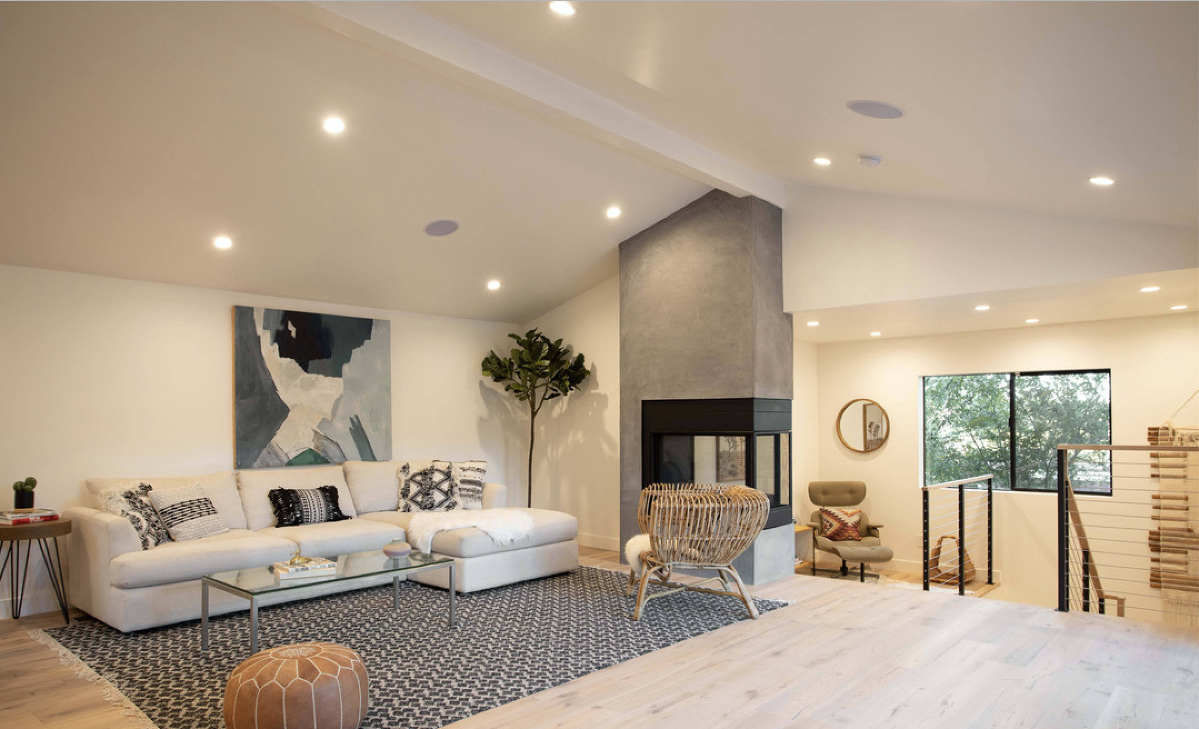 A modern living room features a sofa, a geometric rug, a fireplace, and large windows allowing natural light to enter the space.