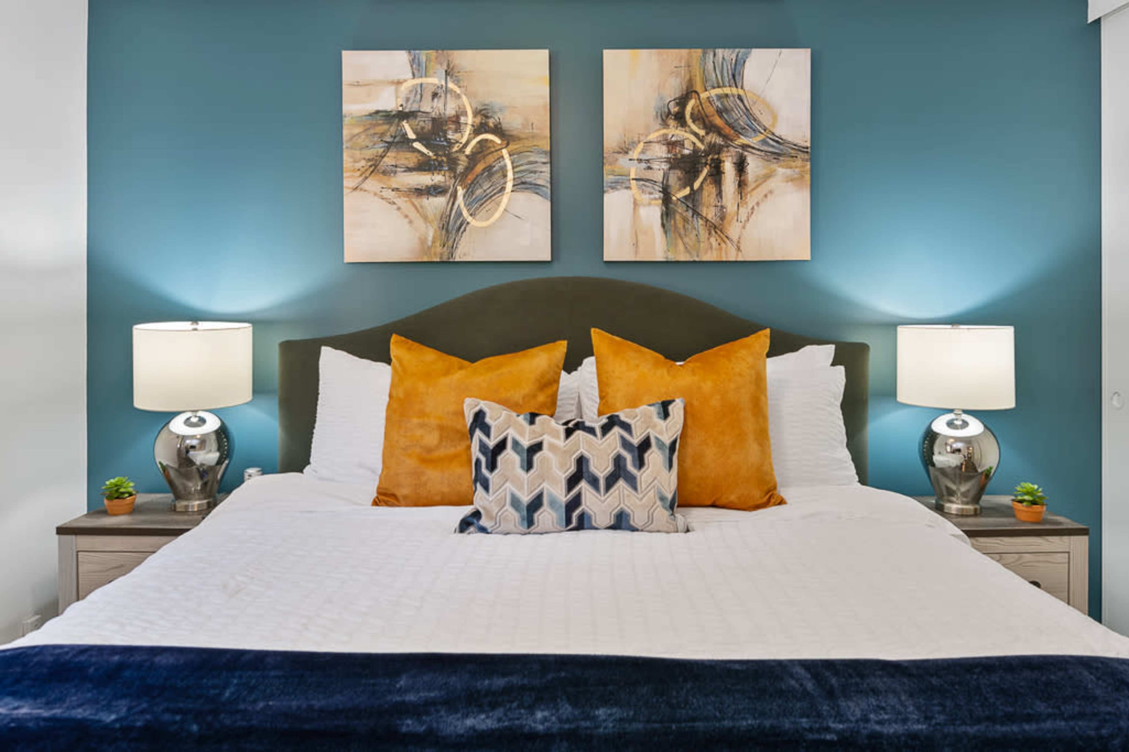 A neatly arranged bedroom features a bed with white linens, two decorative pillows, and abstract artwork above the headboard, flanked by bedside lamps.