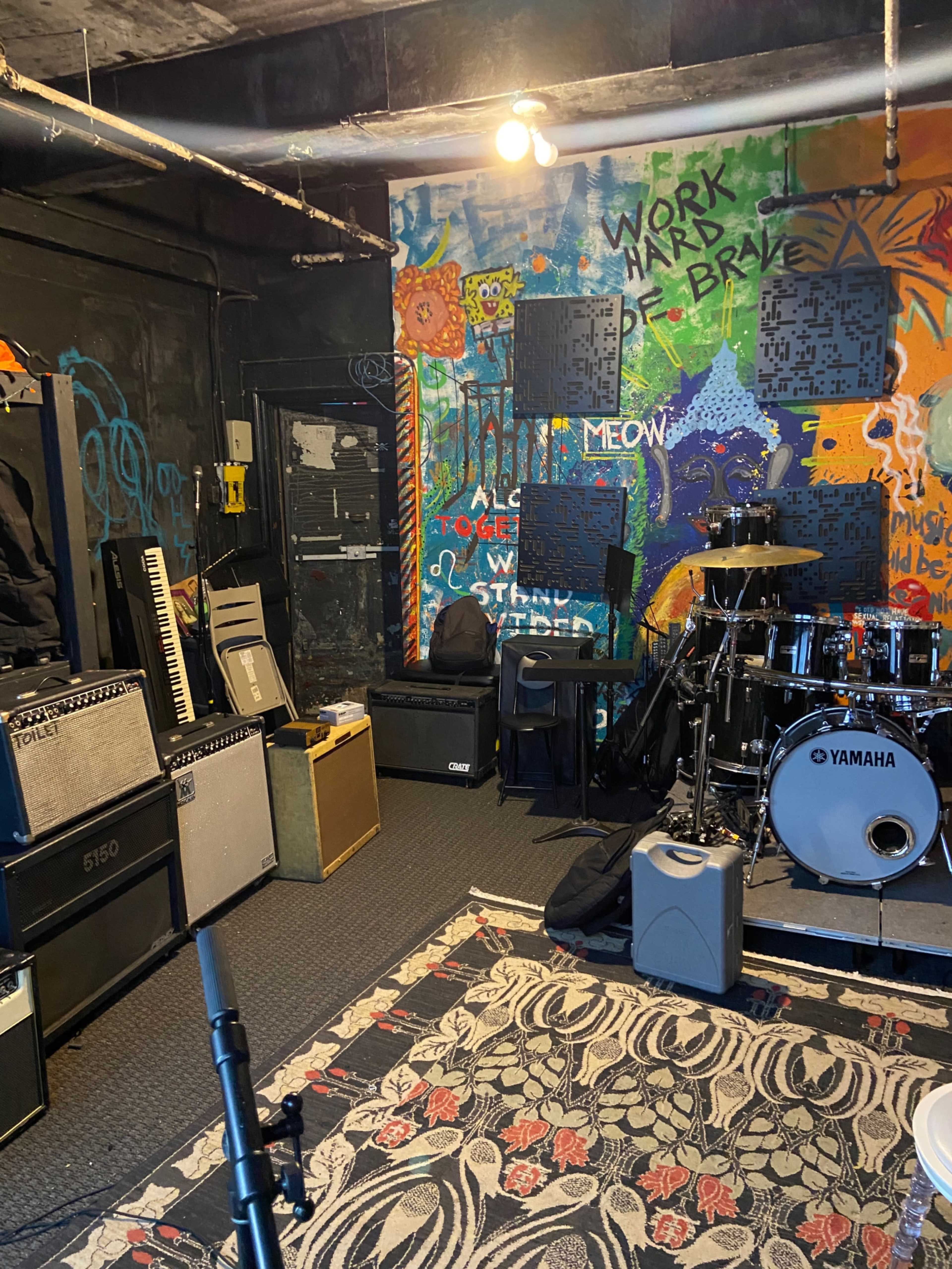 The image shows a music rehearsal space with amplifiers, a drum set, and colorful graffiti on the walls.
