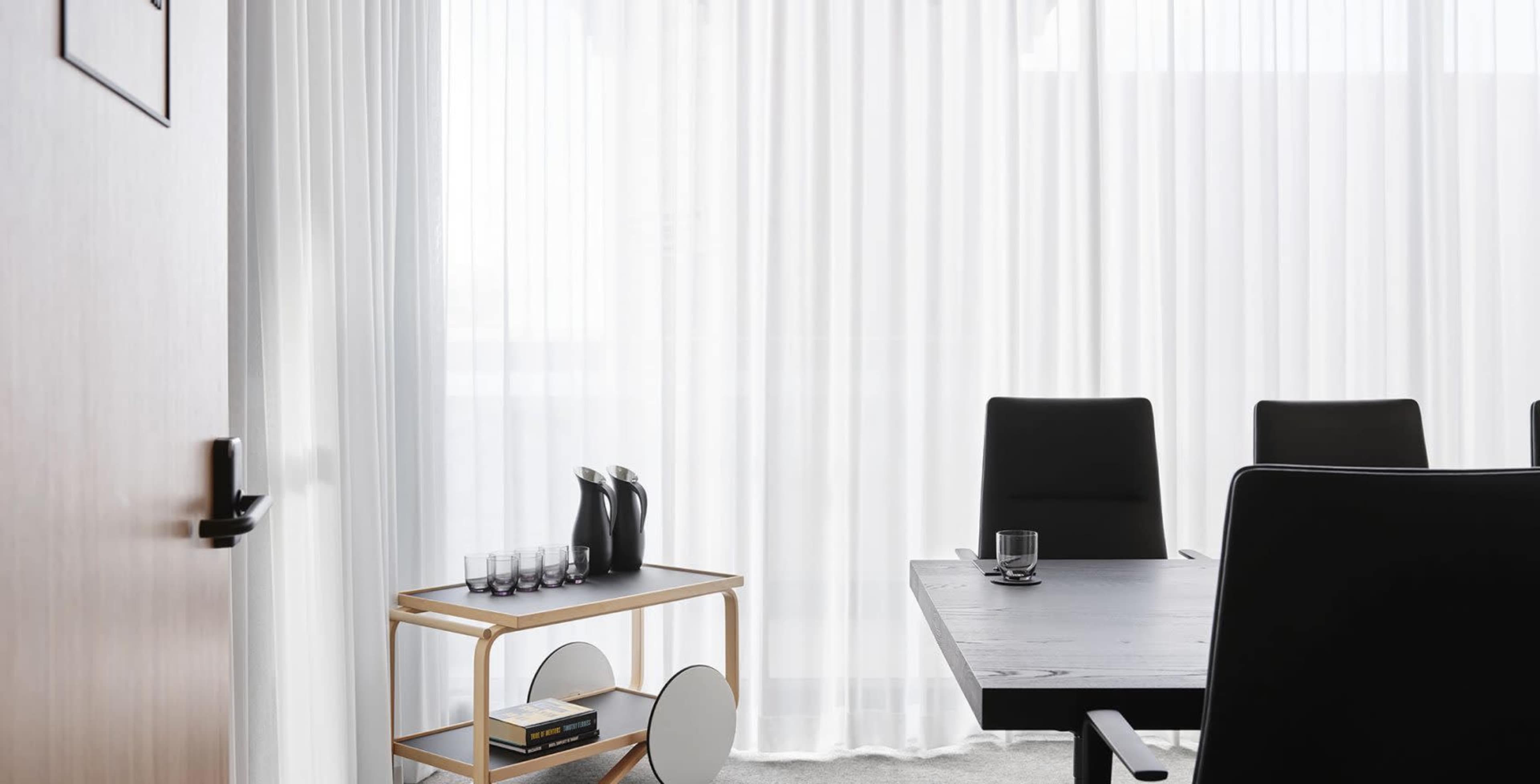 A modern conference room features a large table and black chairs, with a cart displaying glasses and bottles against a backdrop of sheer white curtains.