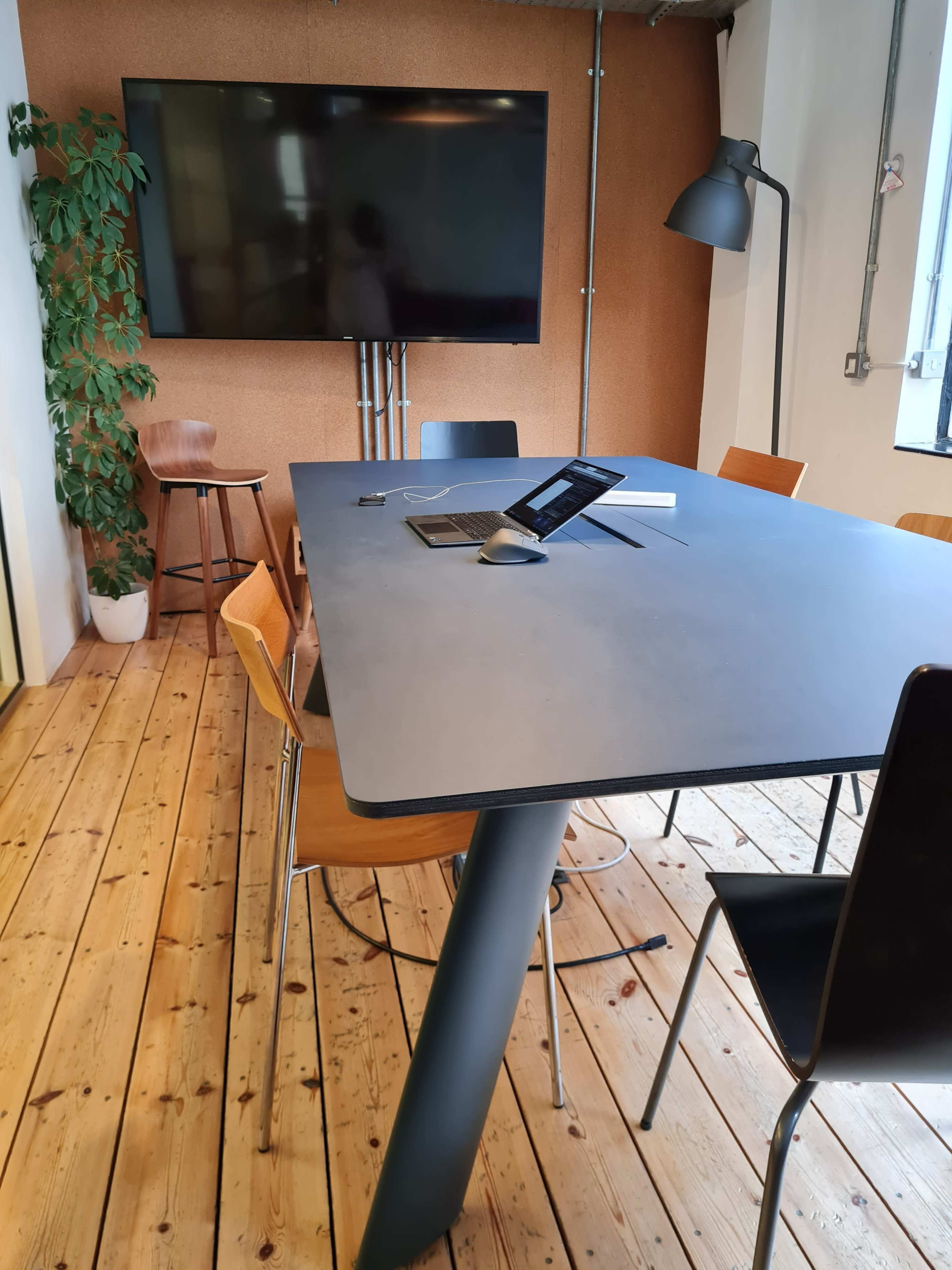 A modern meeting room features a large table surrounded by chairs, a television mounted on the wall, and a plant in the corner.