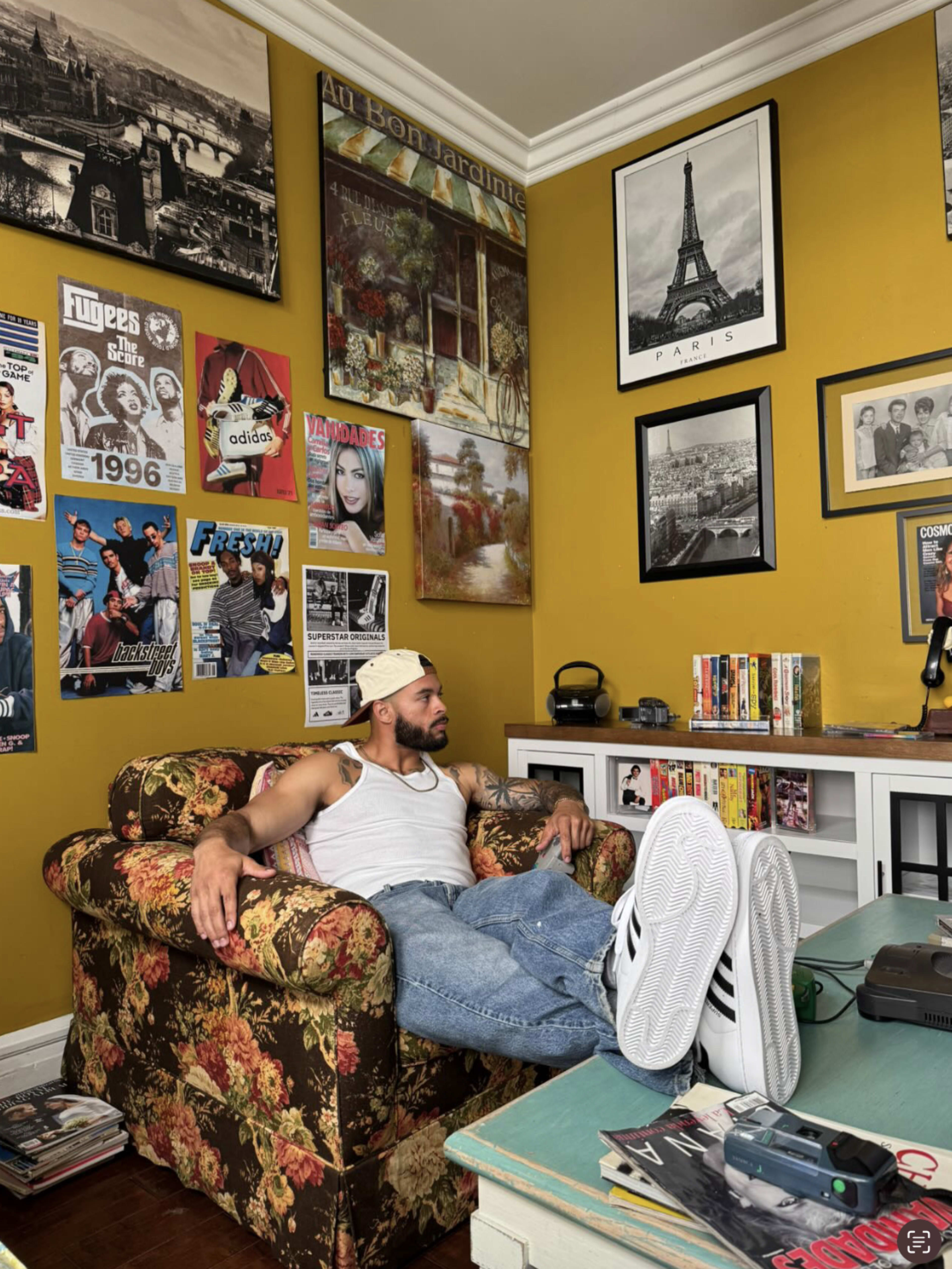 A man in a white tank top and cap sits on a floral couch with his feet up, surrounded by framed artwork and posters on the walls of a yellow room.
