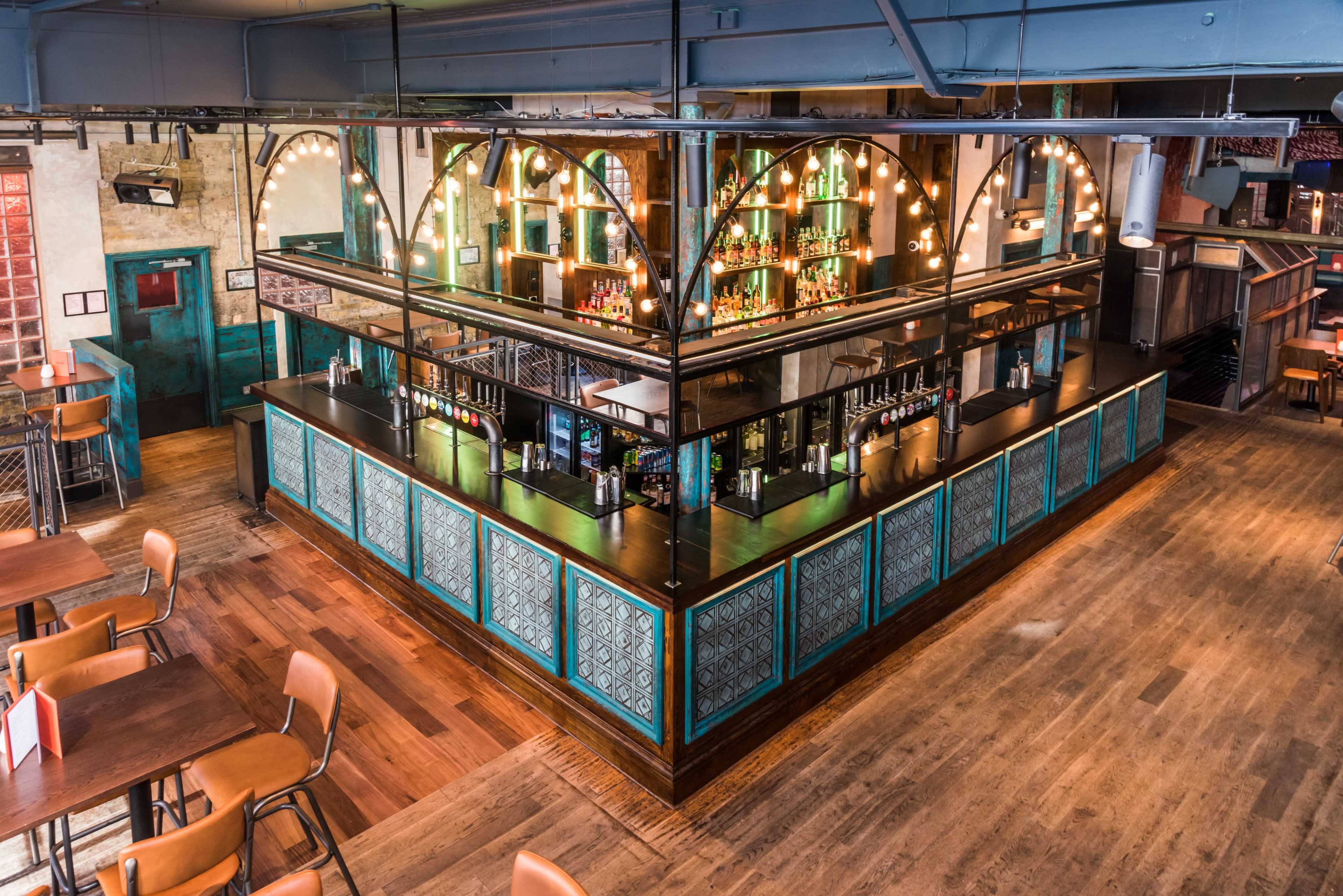 An elevated view of a modern bar area with a glass partition, illuminated shelves, and wooden flooring in a lively restaurant setting.
