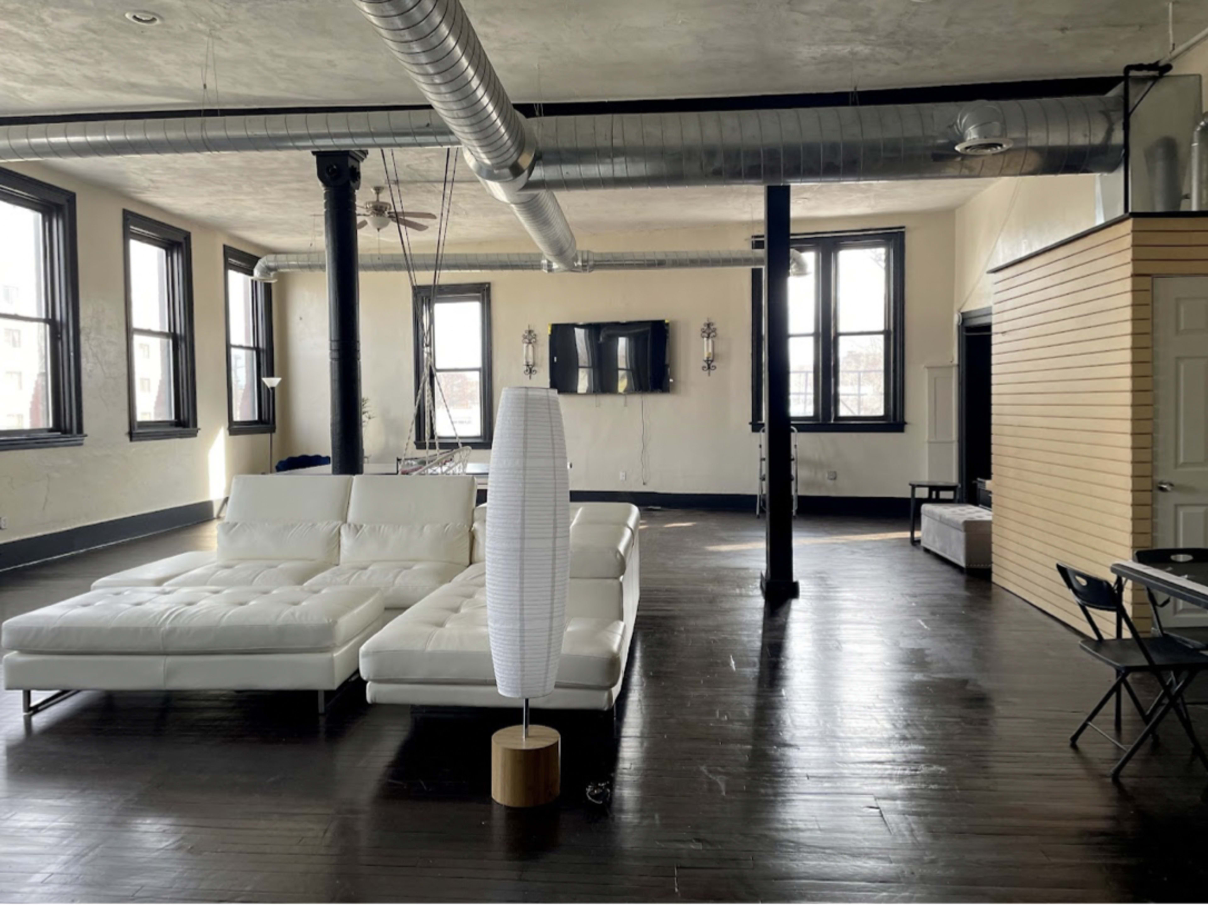 A spacious loft-style living area featuring a white sectional sofa, large windows, and exposed ductwork on the ceiling.