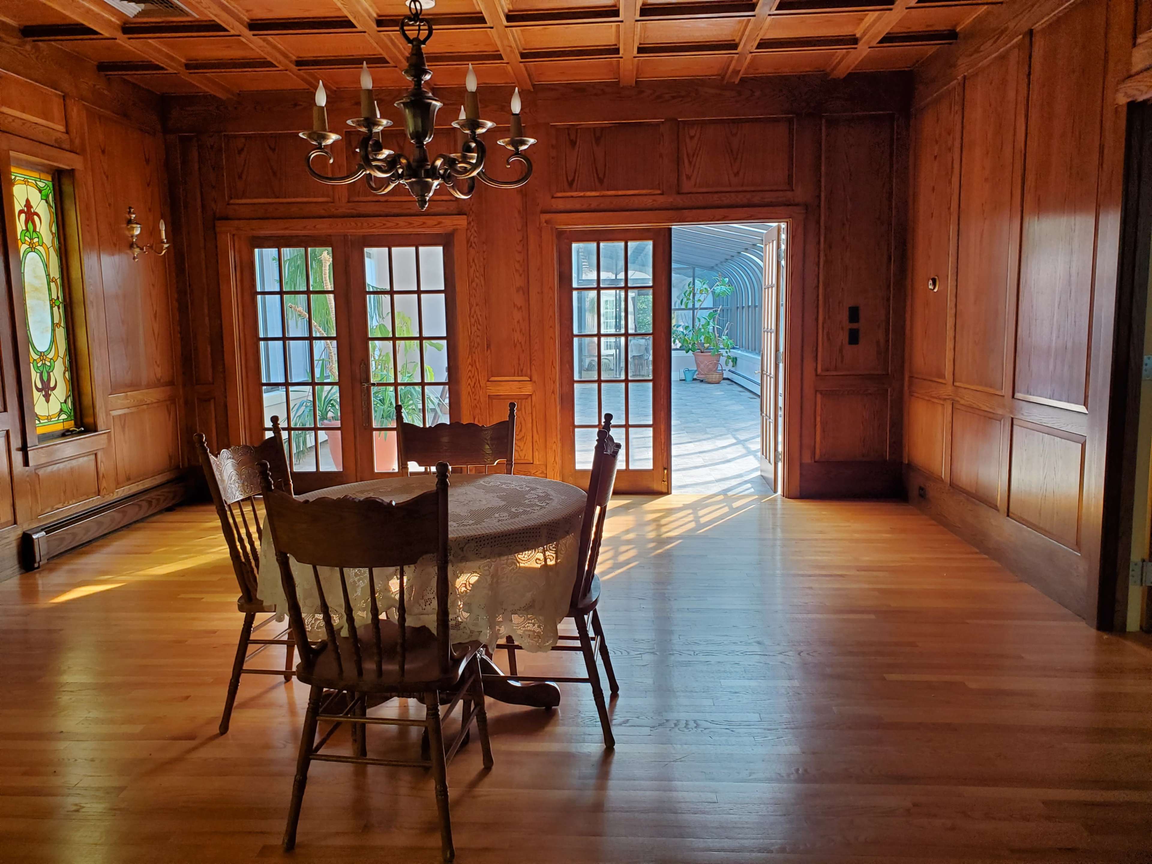 A wooden dining table surrounded by chairs is positioned in a room with paneled walls and large French doors leading to a patio.