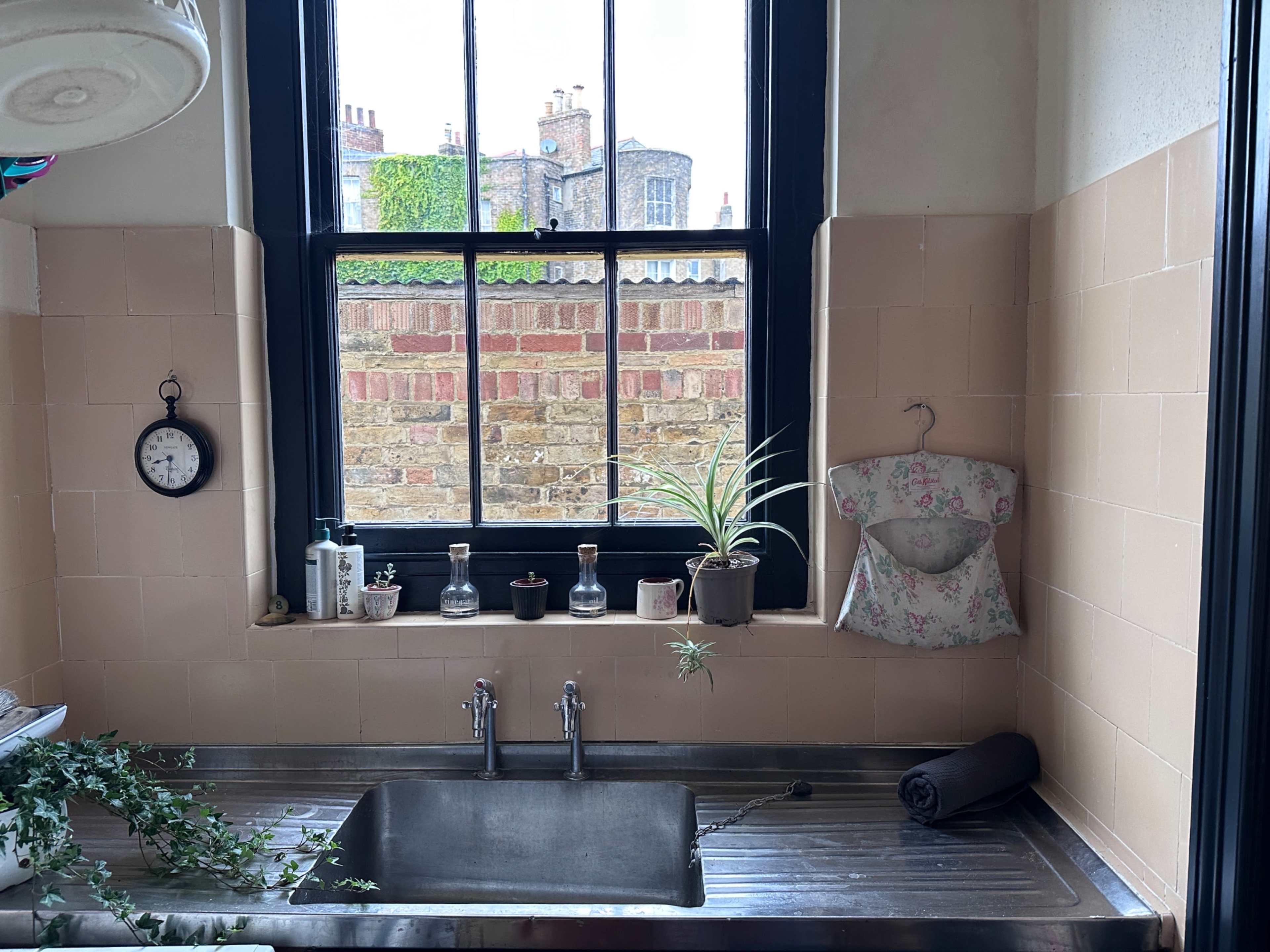 A sink with a stainless steel basin is positioned under a large window, featuring a mix of glass containers, potted plants, and a clock on the surrounding windowsill.