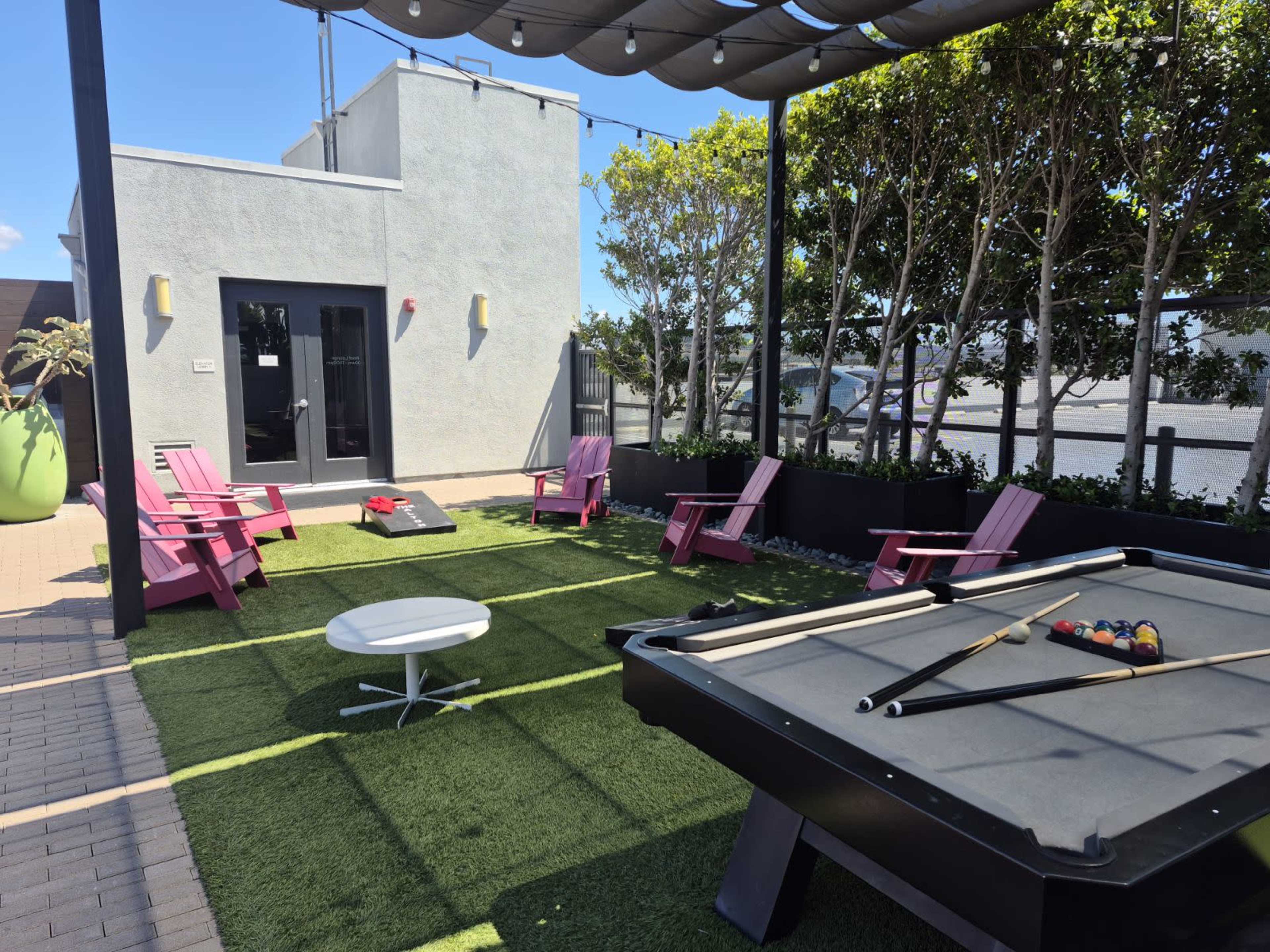 The image depicts an outdoor recreational area featuring a pool table, pink adirondack chairs, a small white table, and artificial grass, surrounded by green trees and a building.