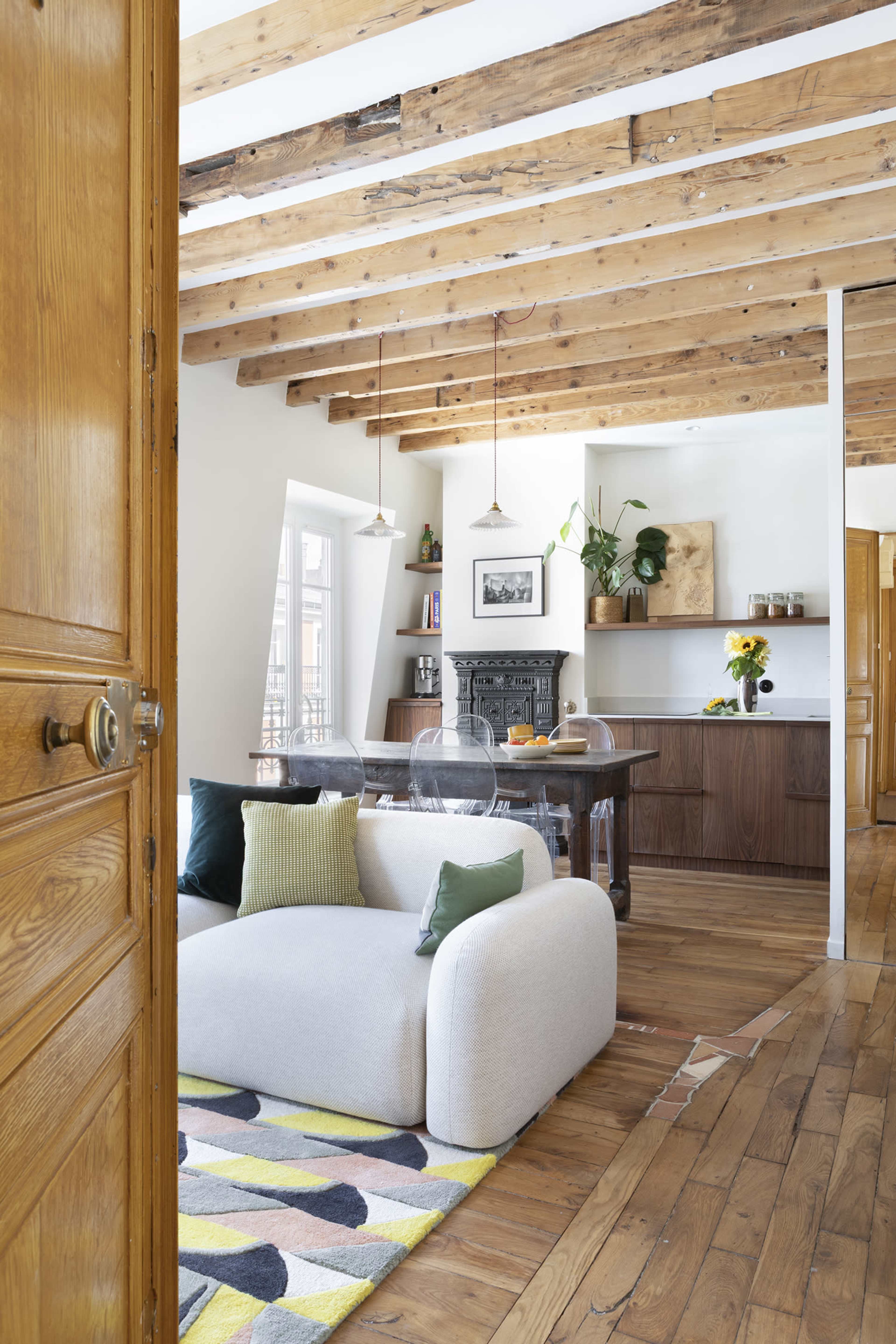 The image shows a modern living space with exposed wooden beams, a light-colored sofa, a dining area with a wooden table, and an open layout featuring a kitchen and decorative plants.
