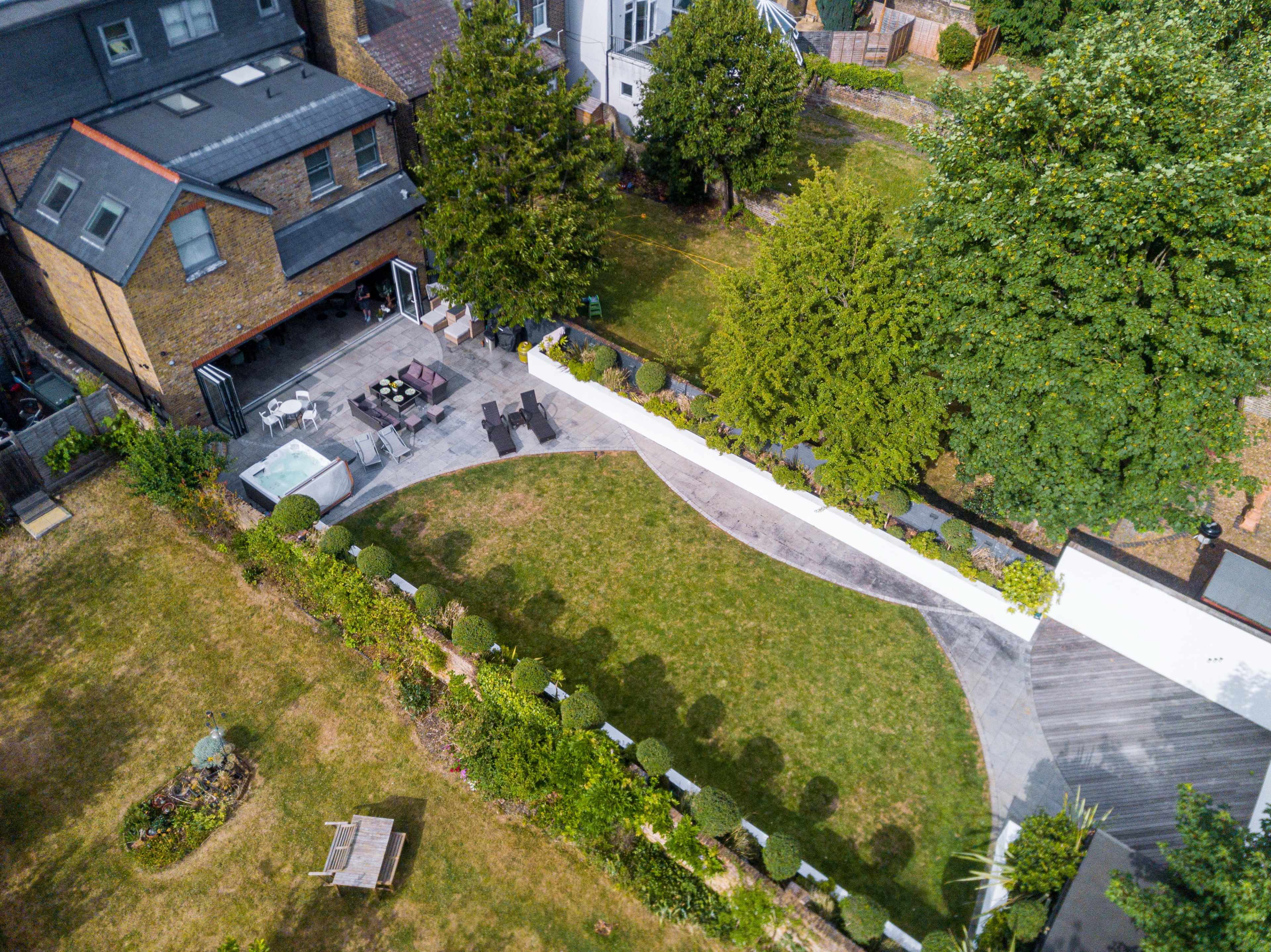 An aerial view shows a landscaped garden with a patio area, seating, and a hot tub, surrounded by greenery and residential buildings.