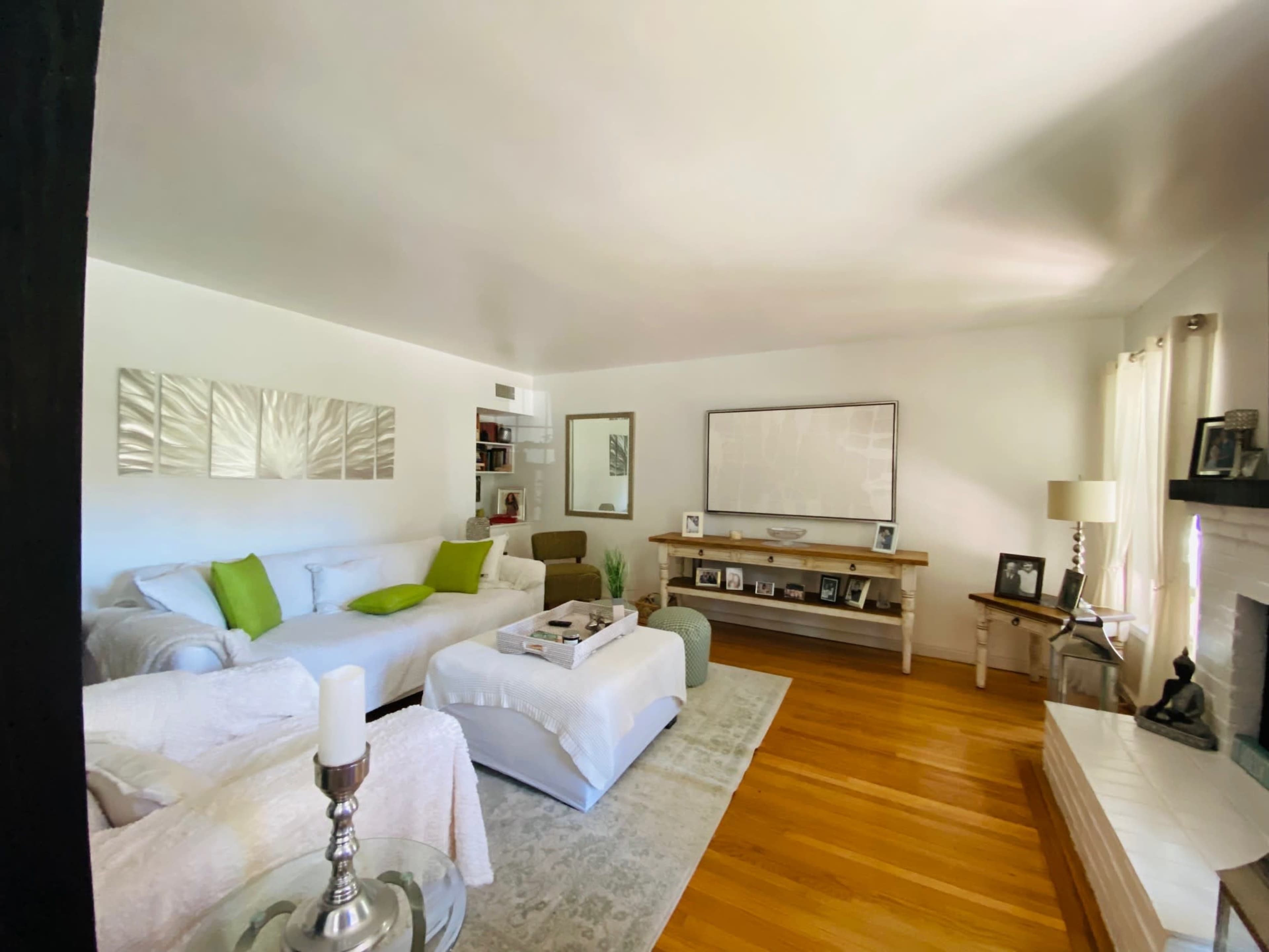 The living room features white sofas, a wooden shelf with photographs, and a large abstract wall art piece centered above a light wooden table.