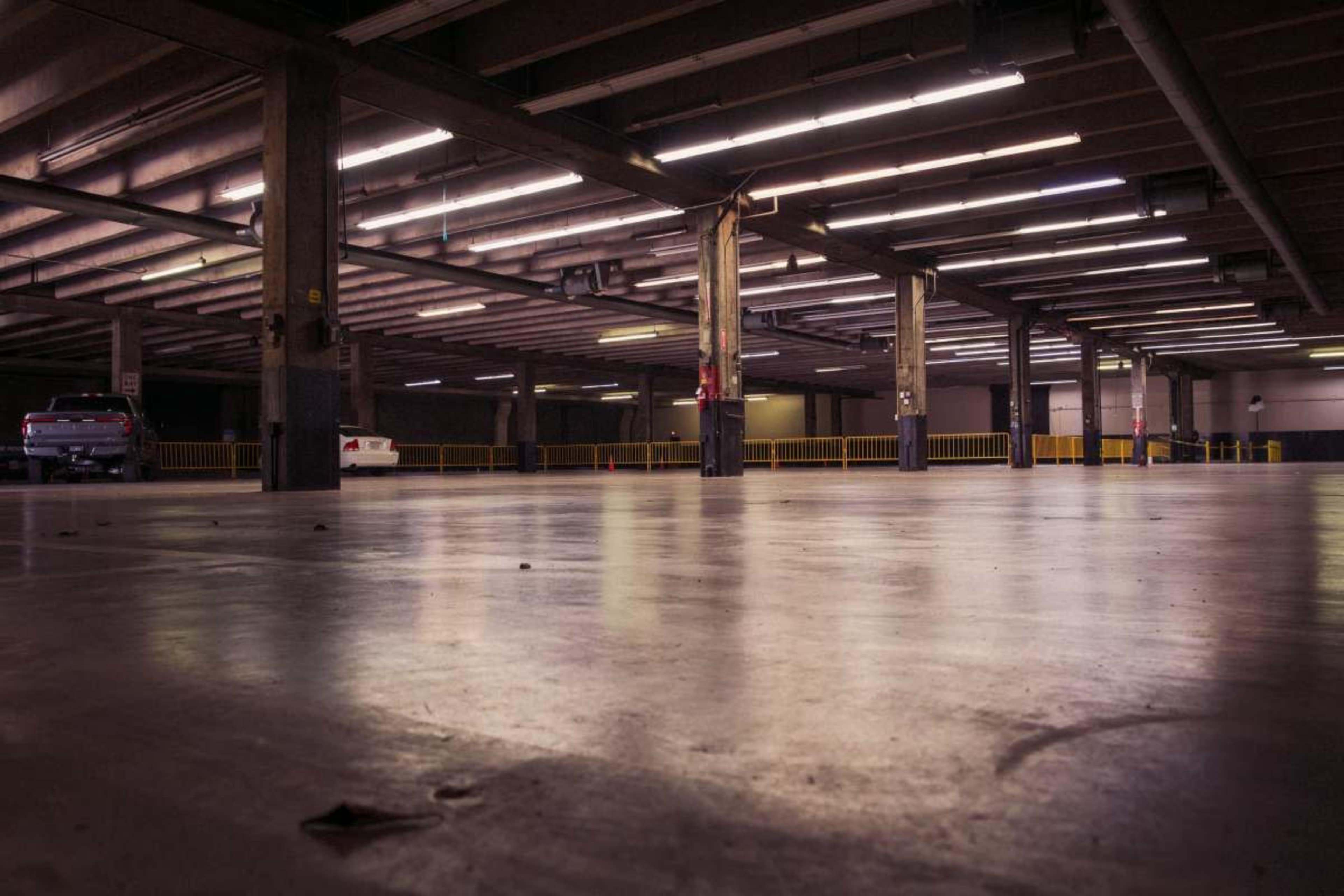 A dimly lit, empty parking garage features concrete flooring and several parked vehicles.