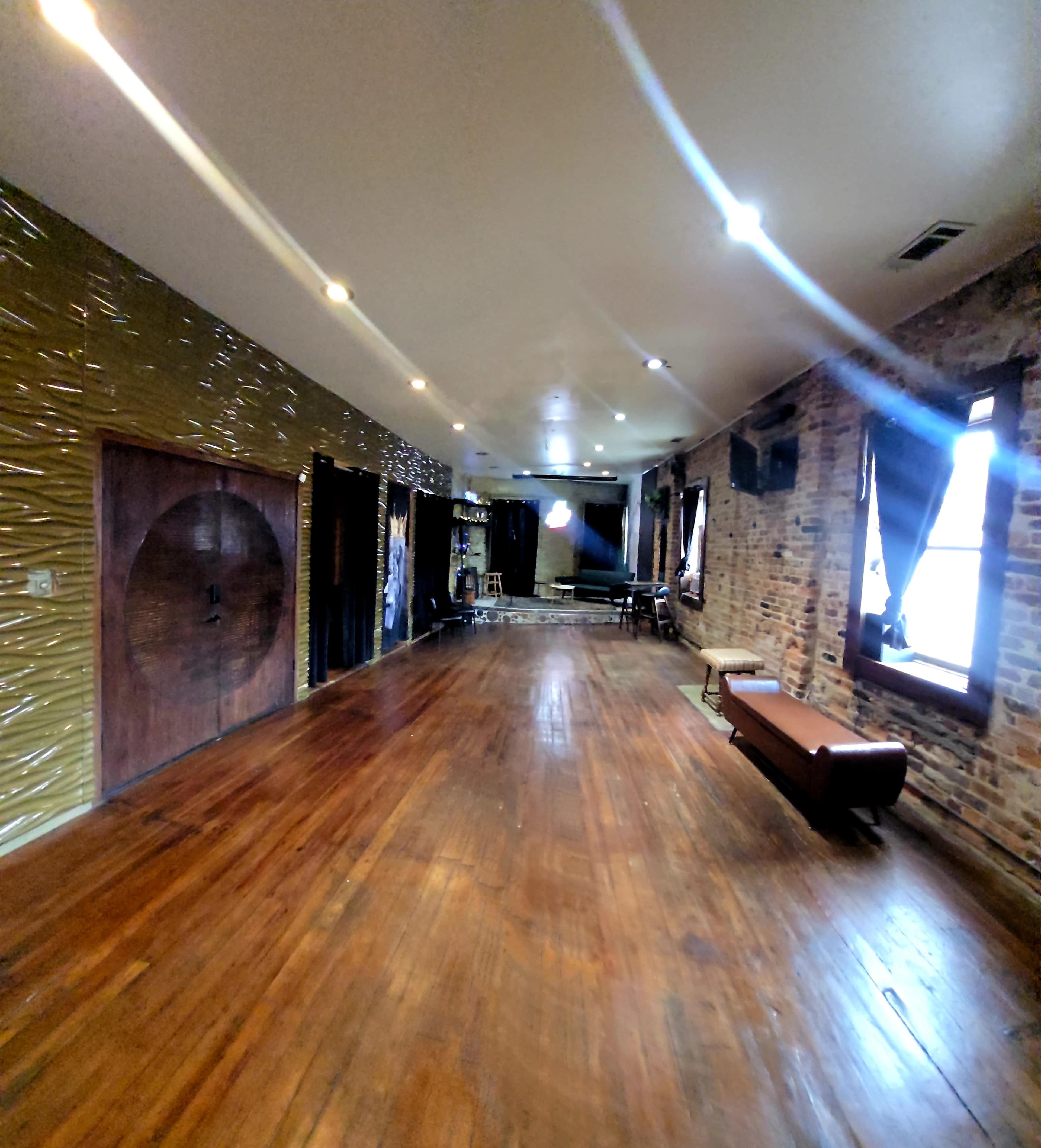 The image shows a spacious room with wooden floors, brick walls, and various lighting fixtures, featuring a bench and a door in the background.