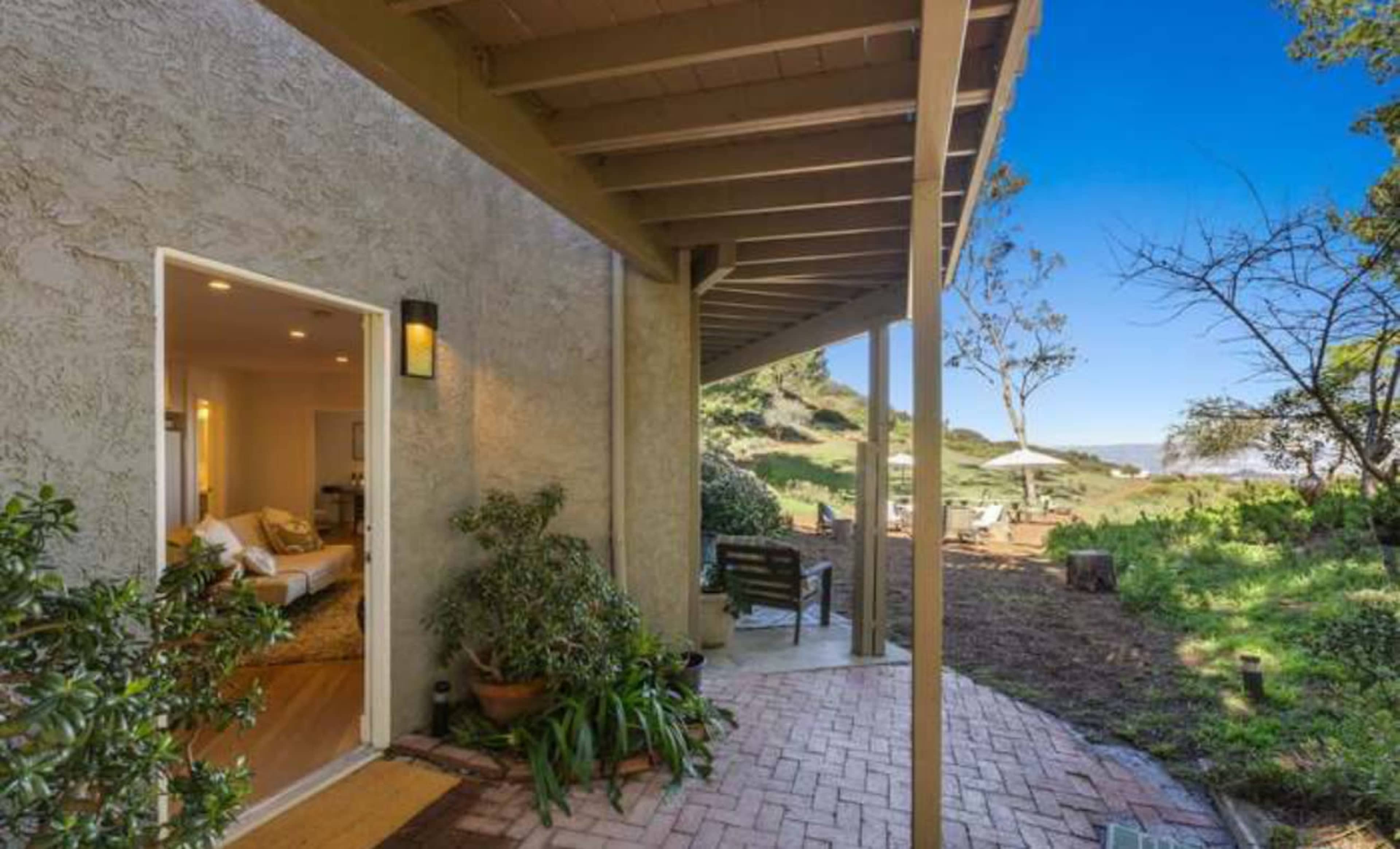 A pathway leads from an entrance of a house to a garden area with outdoor seating and a view of the landscape.