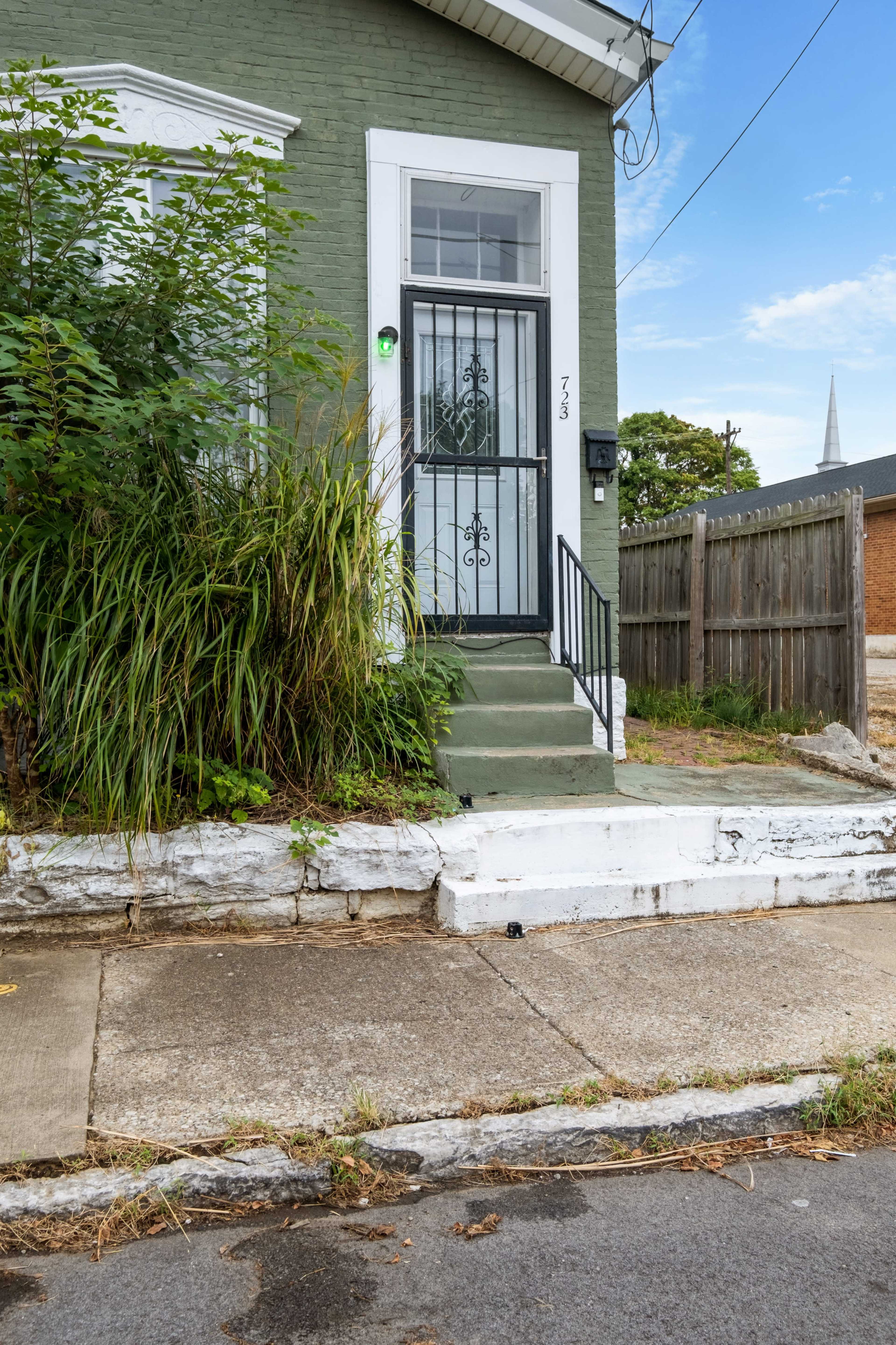 A green house with a wrought iron door and overgrown vegetation at the entrance.