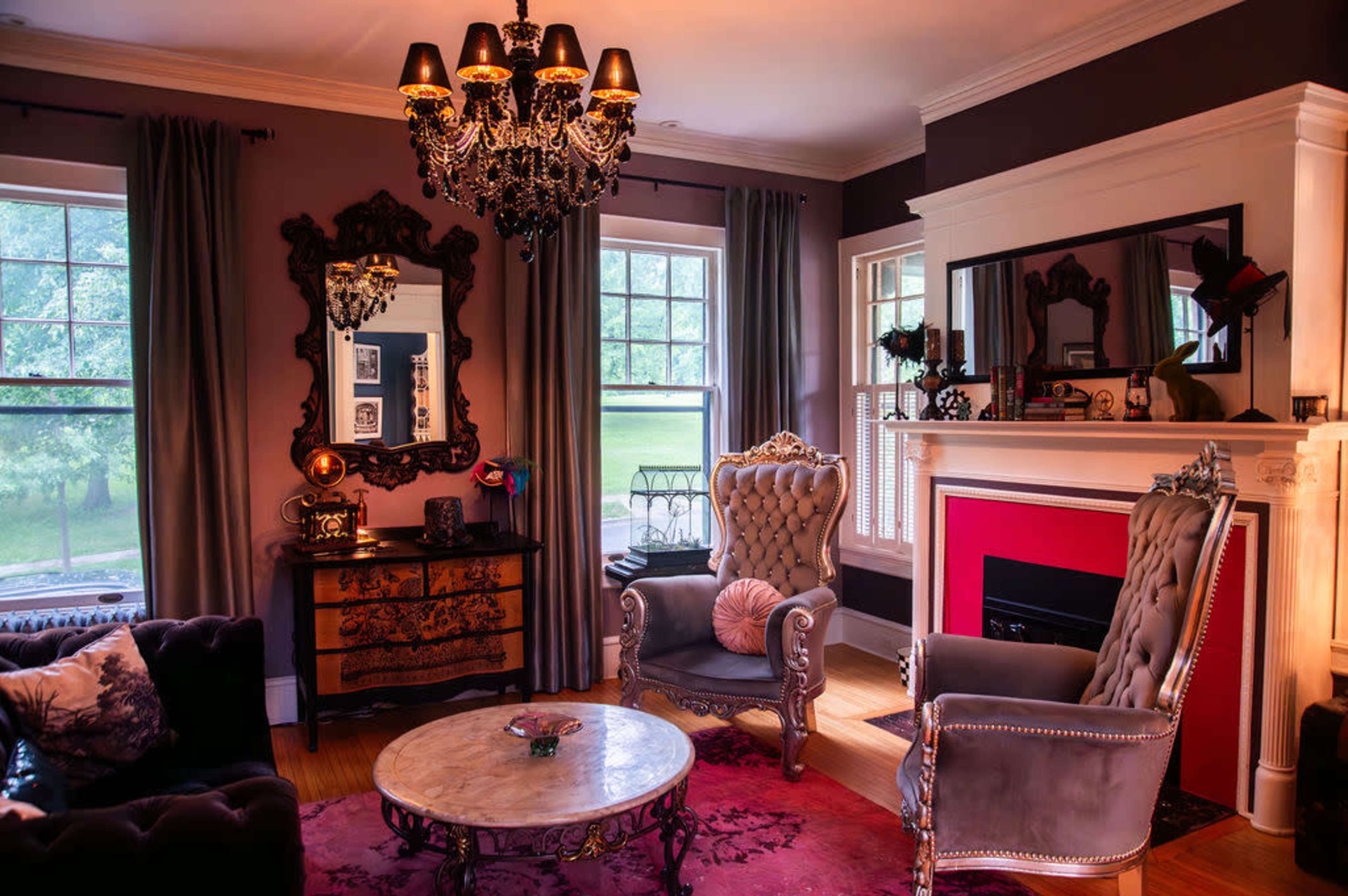 The image shows a stylish living room with ornate furniture, a chandelier, and large windows overlooking a green outdoor area.