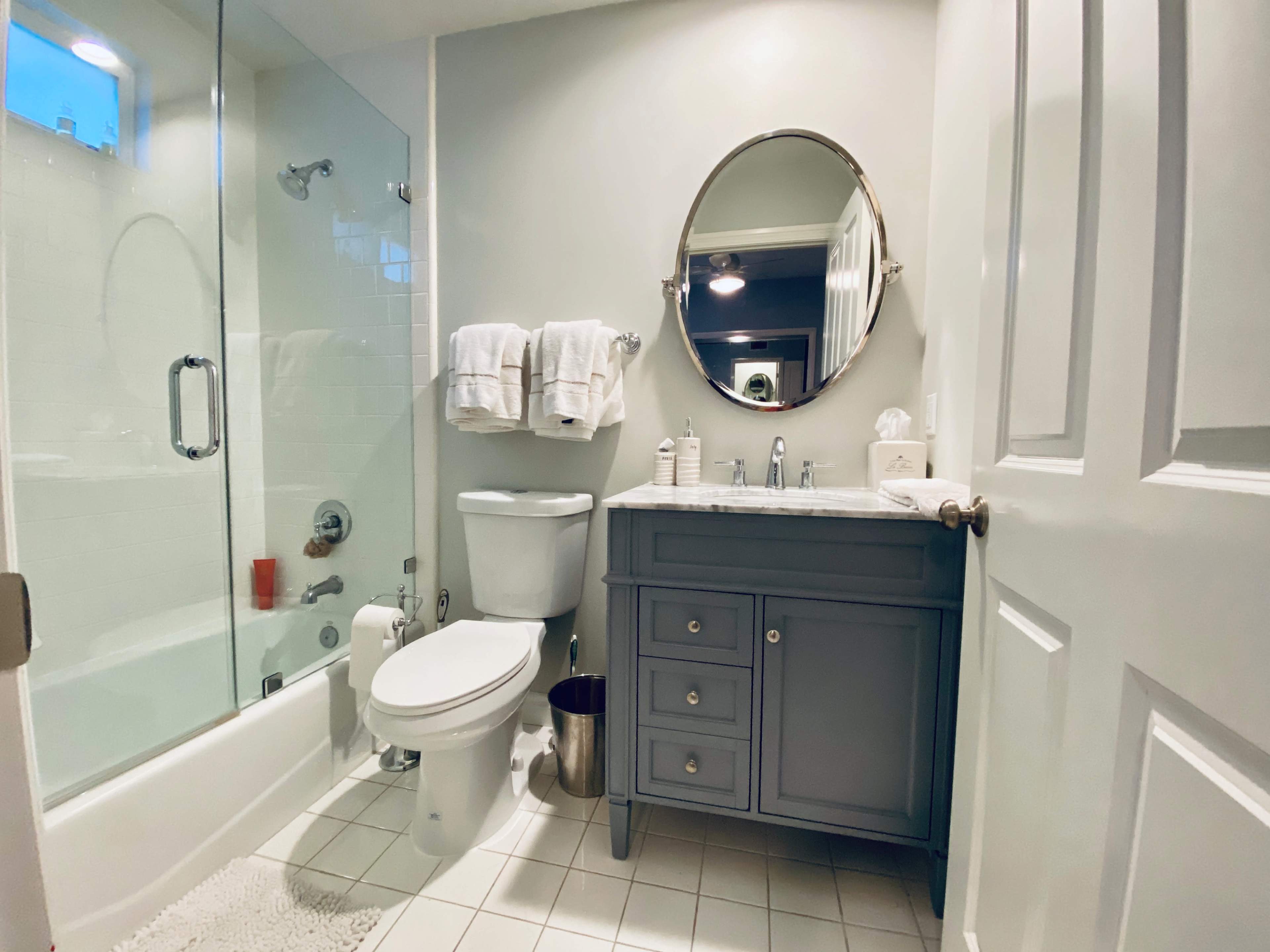 A modern bathroom with a glass-enclosed shower, toilet, and a gray vanity with a circular mirror above it.